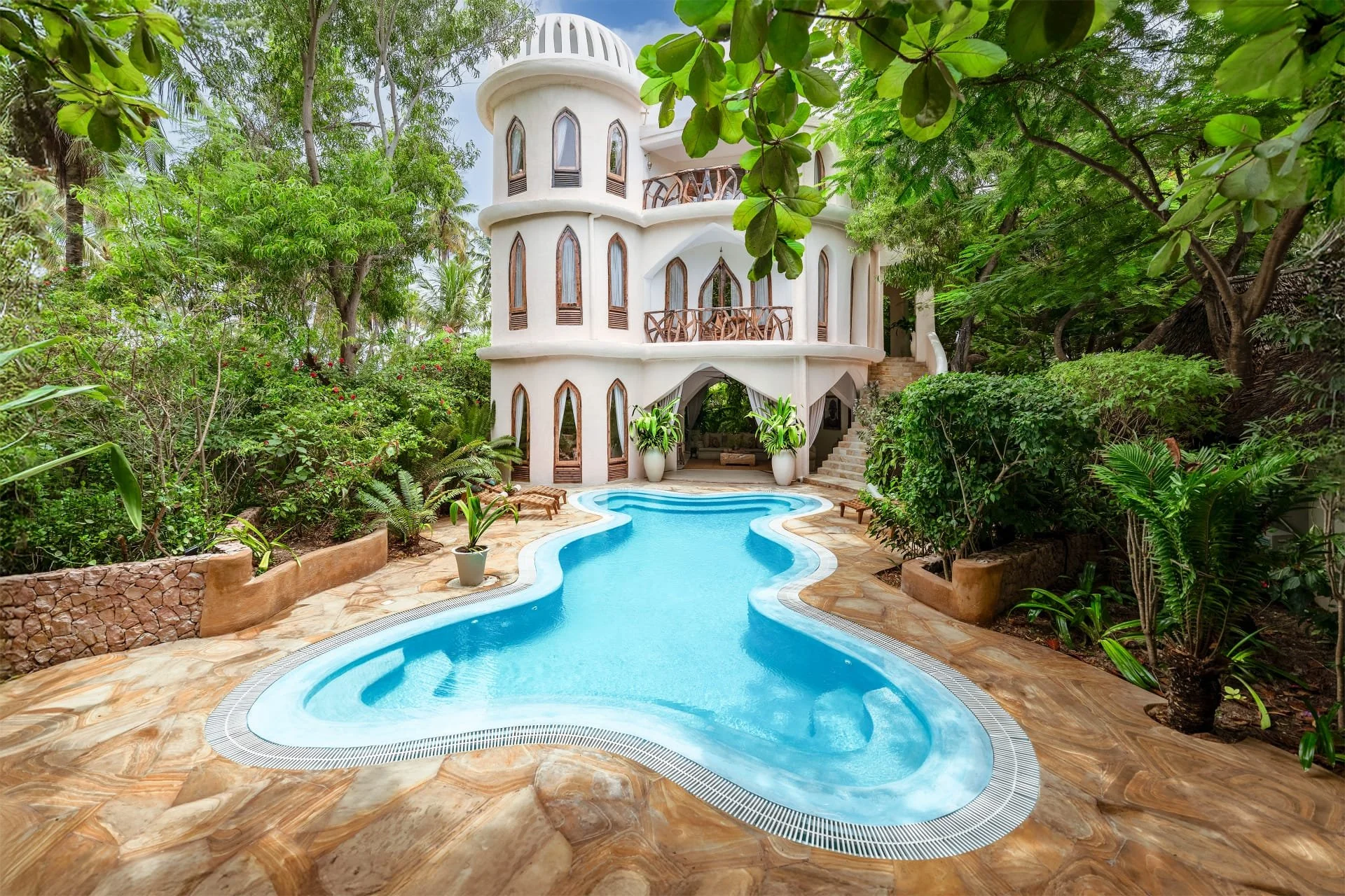 Ein tropischer Garten mit einem kurvenförmigen Swimmingpool vor einem weißen, mehrstöckigen Haus mit Rundbalkonen und hohen, schmalen Fenstern, umgeben von üppigem Grün und Bäumen.