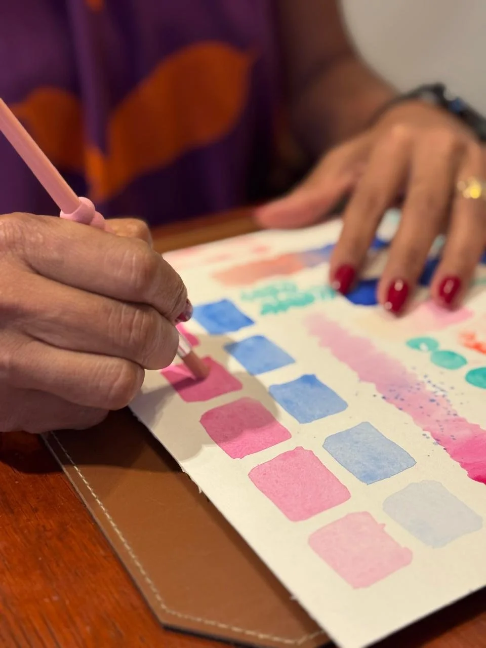 Close-up of a person writing or drawing on colorful paper with pastel pink and blue squares, using a pink pen. The person has painted nails and is wearing a ring.