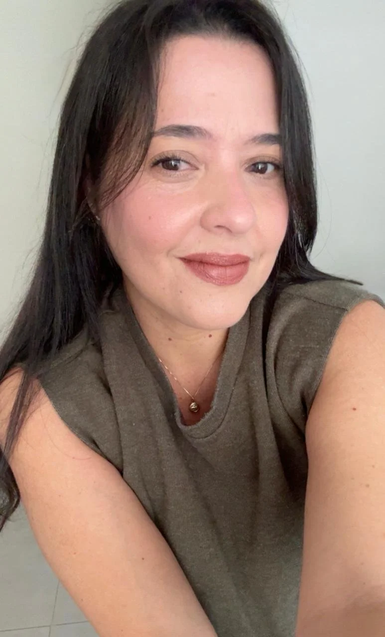 A woman taking a selfie with a slight smile, wearing a brown sleeveless top and a gold necklace, with straight dark hair falling over her shoulders.