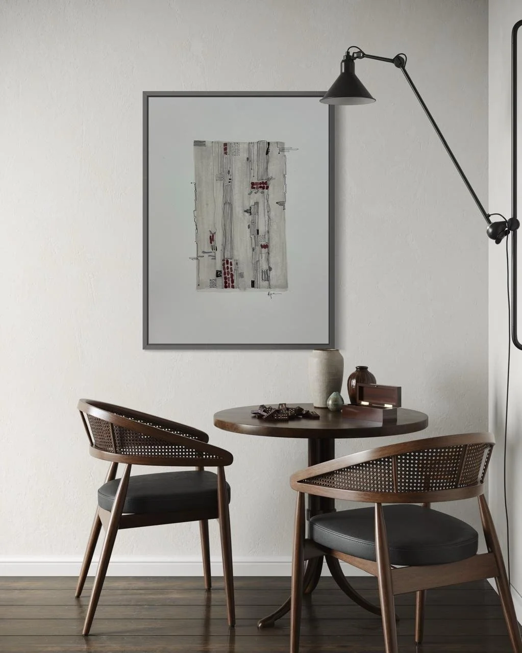 A round wooden table with two matching wood chairs with black cushioned seats. The table has decorative vases, a small box, and a decorative object. A framed abstract artwork hangs on the white wall above, and a black adjustable wall lamp is mounted next to the artwork.
