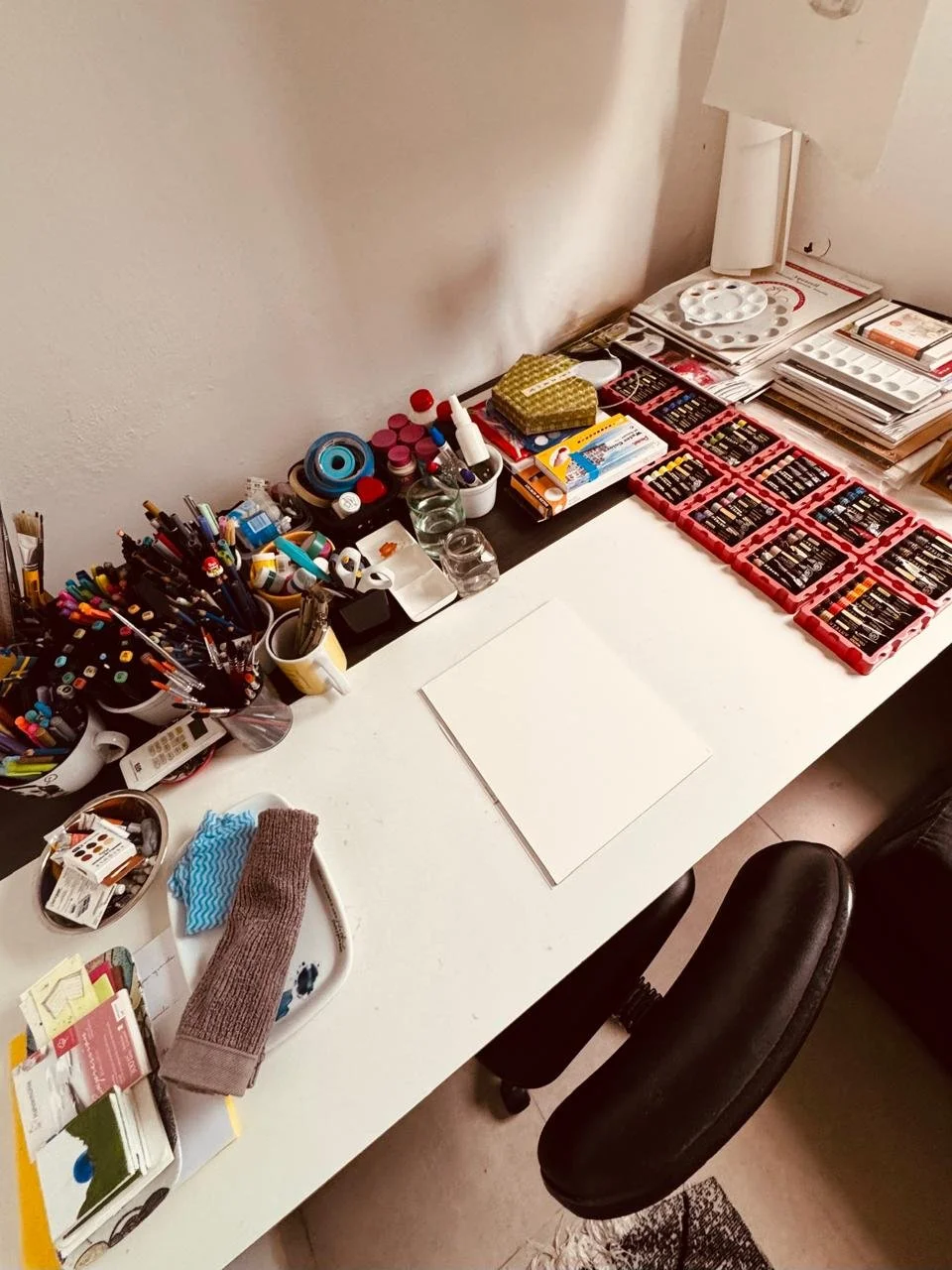 Overhead view of a cluttered desk with pens, bottles, and office supplies, alongside art supplies including paints, brushes, and paper, in a home workspace.