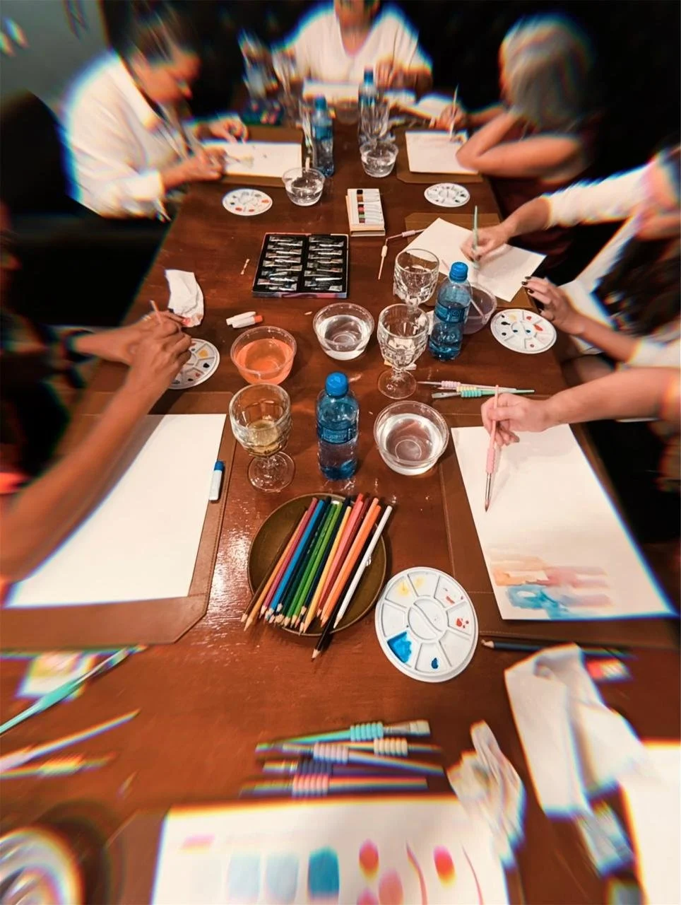 People gathered around a wooden table engaged in a painting or art activity, with brushes, paints, water bottles, and glasses of water on the table.