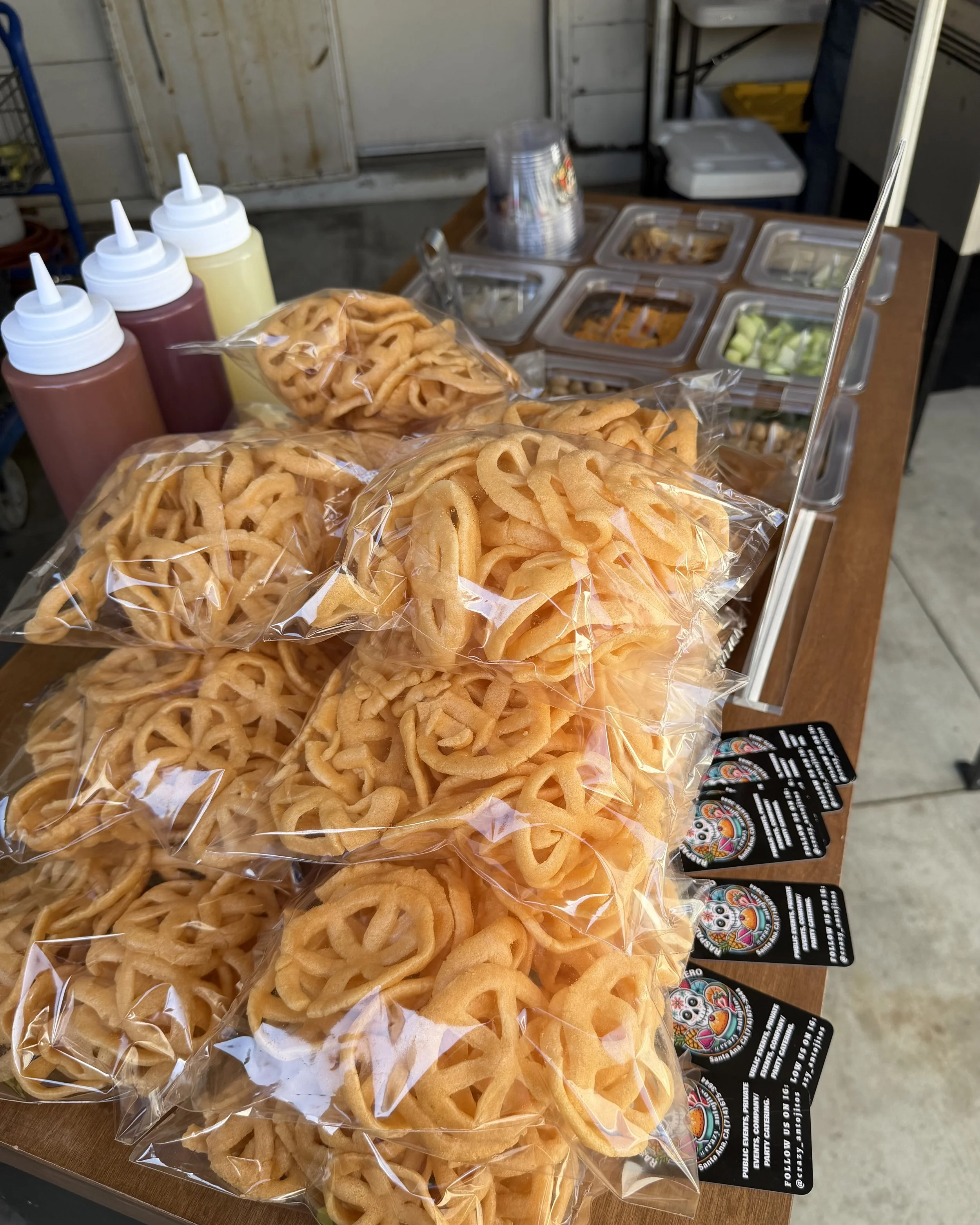 Fried onion rings in clear plastic bags on a table with squeeze bottles of ketchup, mustard, and other condiments, and containers of toppings such as chopped green onions and sauces in the background.