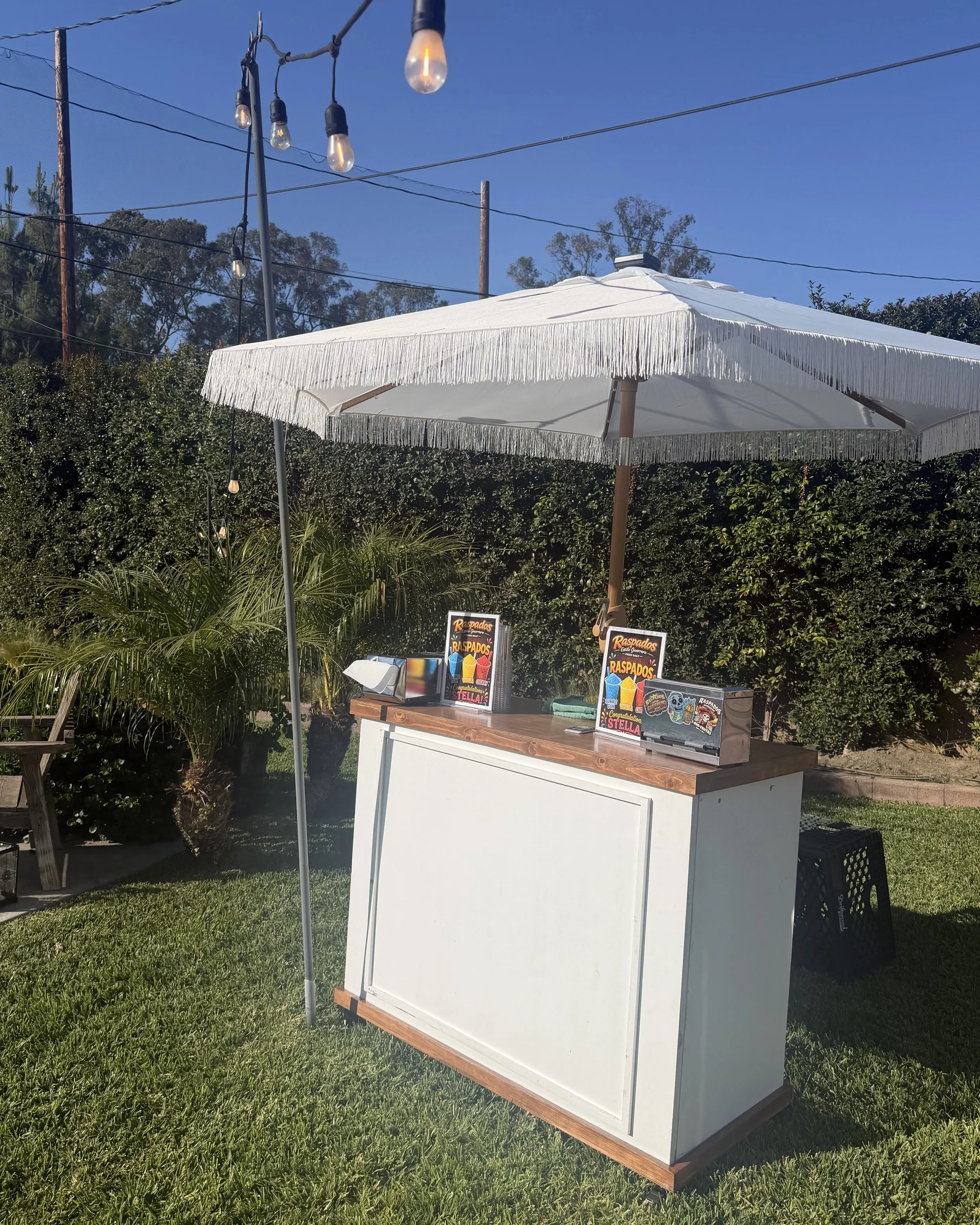Outdoor scene with a white bar or counter with colorful menu signs, under a large white umbrella with fringe, surrounded by greenery, outdoor string lights, and a blue sky.