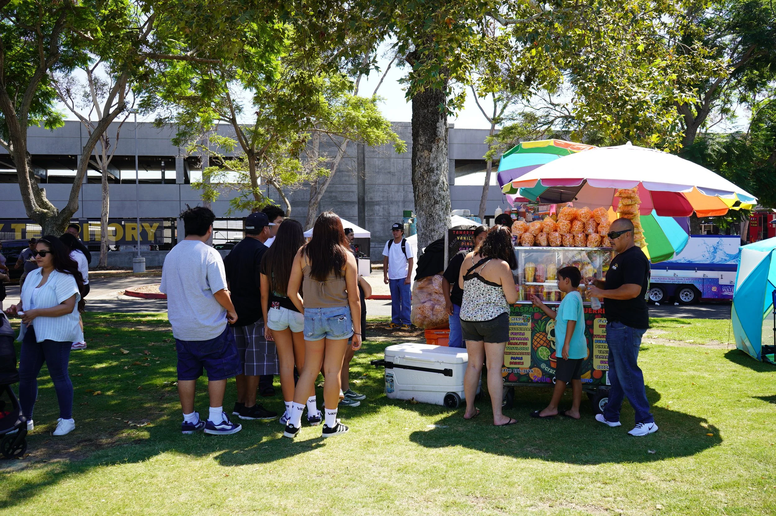 People gathered near a colorful street food cart with umbrellas in a park, surrounded by trees and a concrete building in the background.