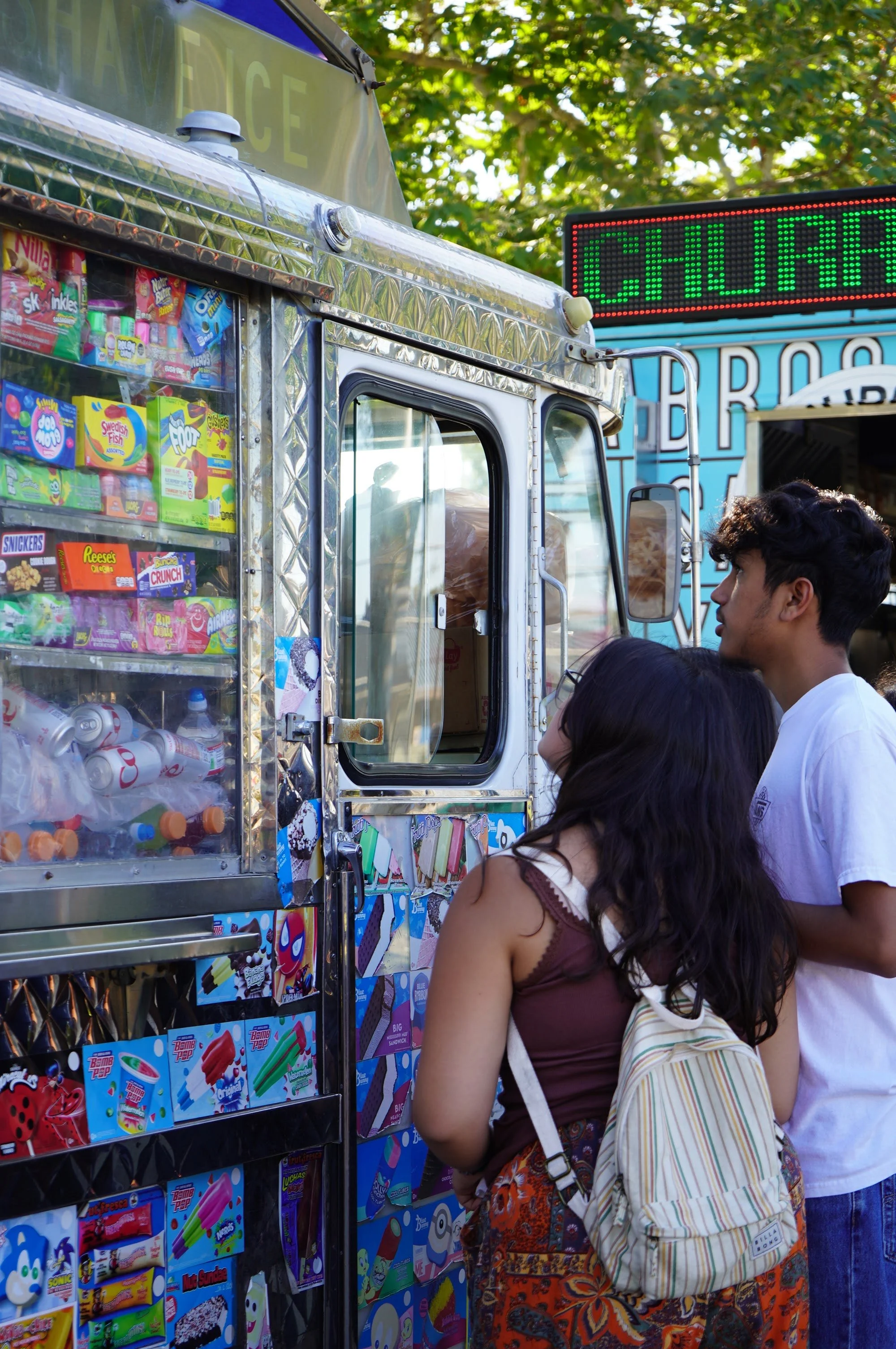 People in line at a colorful food truck selling snacks and ice cream at an outdoor event, with trees in the background.