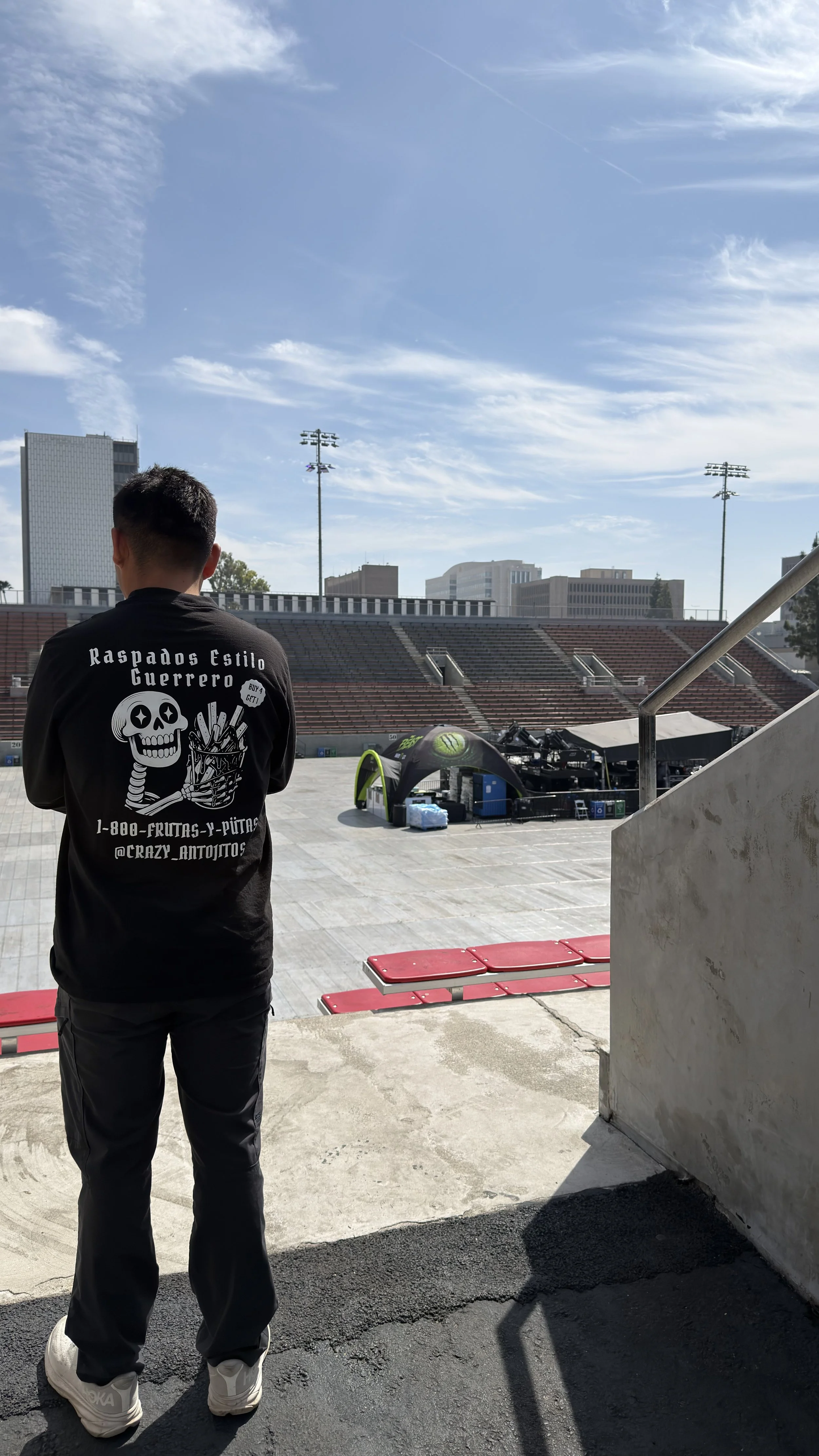 A man stands outdoors in front of a stadium, wearing a black hoodie with a skull and fireworks graphic on the back and some text, with city buildings and blue sky with clouds in the background.