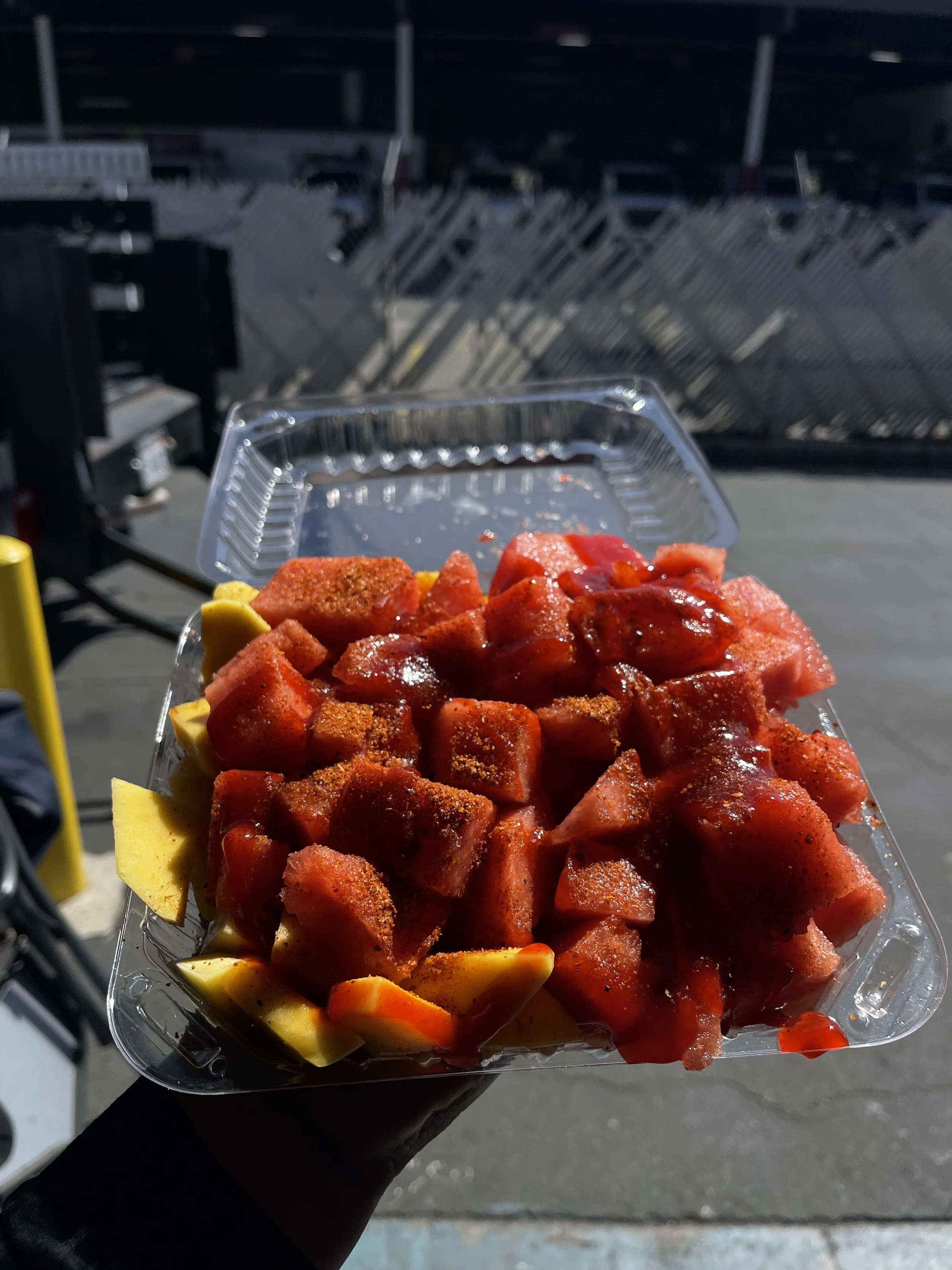A clear plastic container of diced watermelon and a lemon wedge, held by a person outdoors with a dark background.