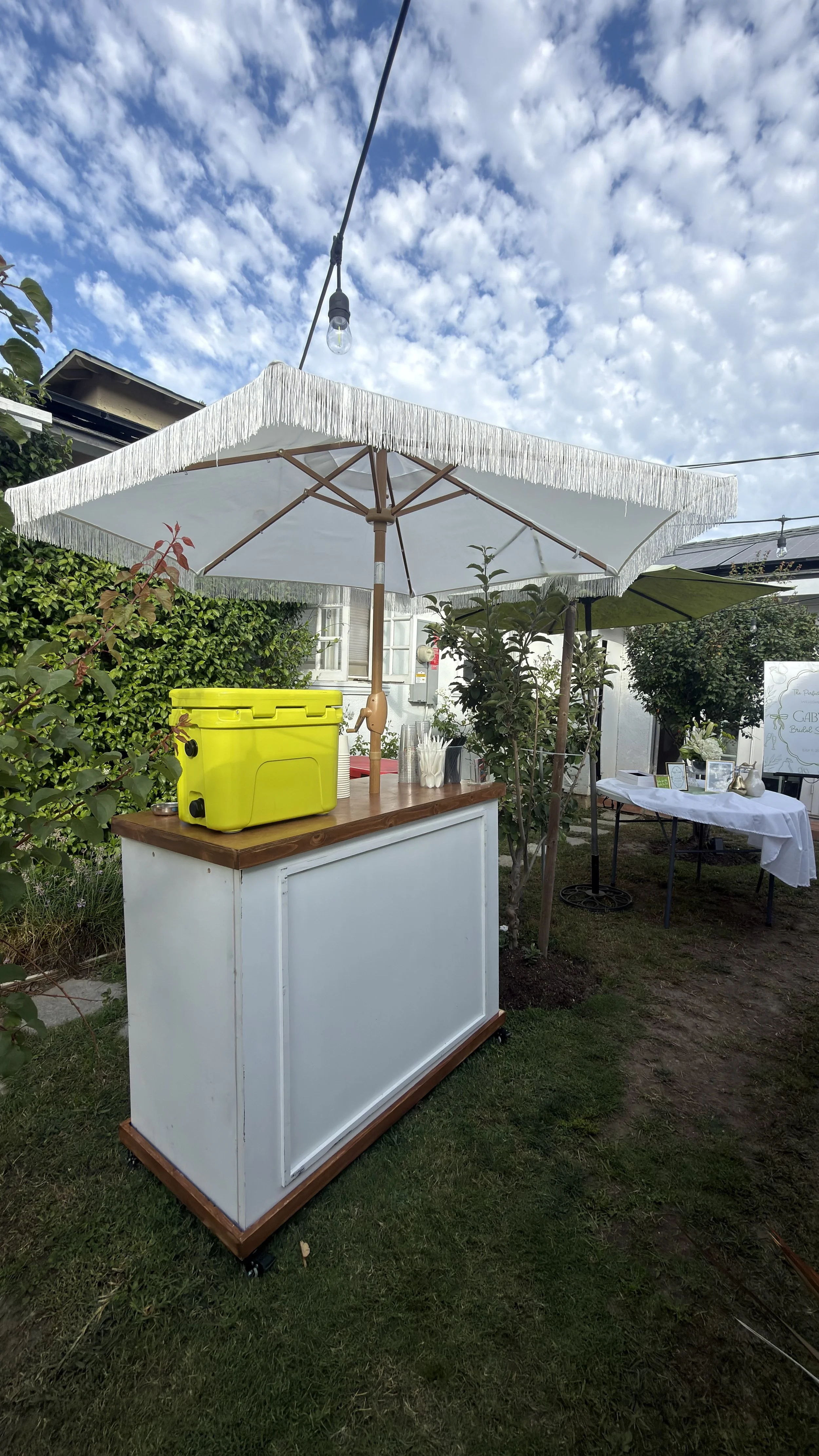 An outdoor mobile bar with a white panel and wooden accents, set under a large white fringed parasol, with a yellow cooler on top, surrounded by greenery and additional tables in the background.