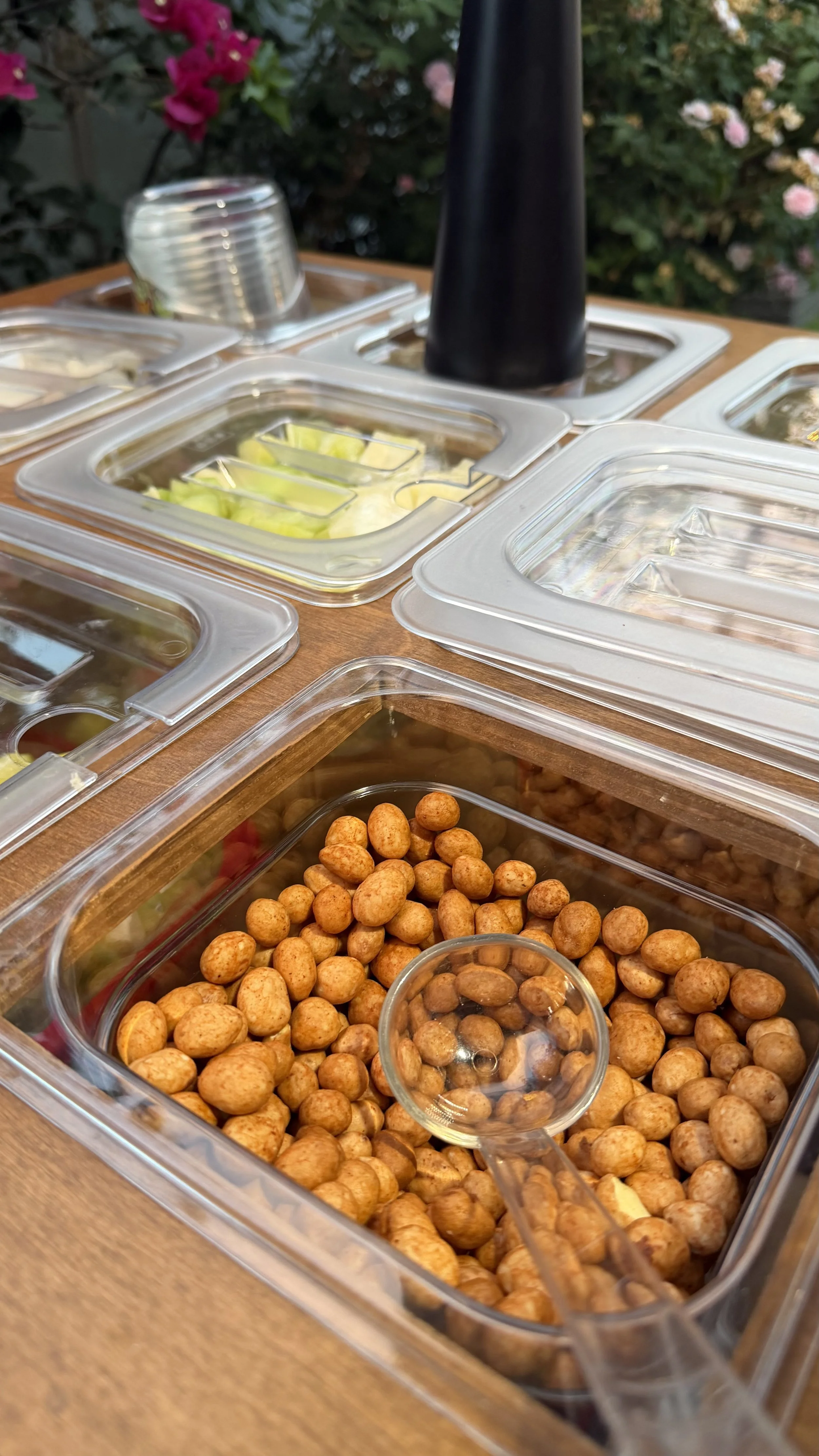 Assorted toppings in clear plastic containers, including roasted chickpeas, sliced cucumbers, and other vegetables, on a wooden table outdoors.