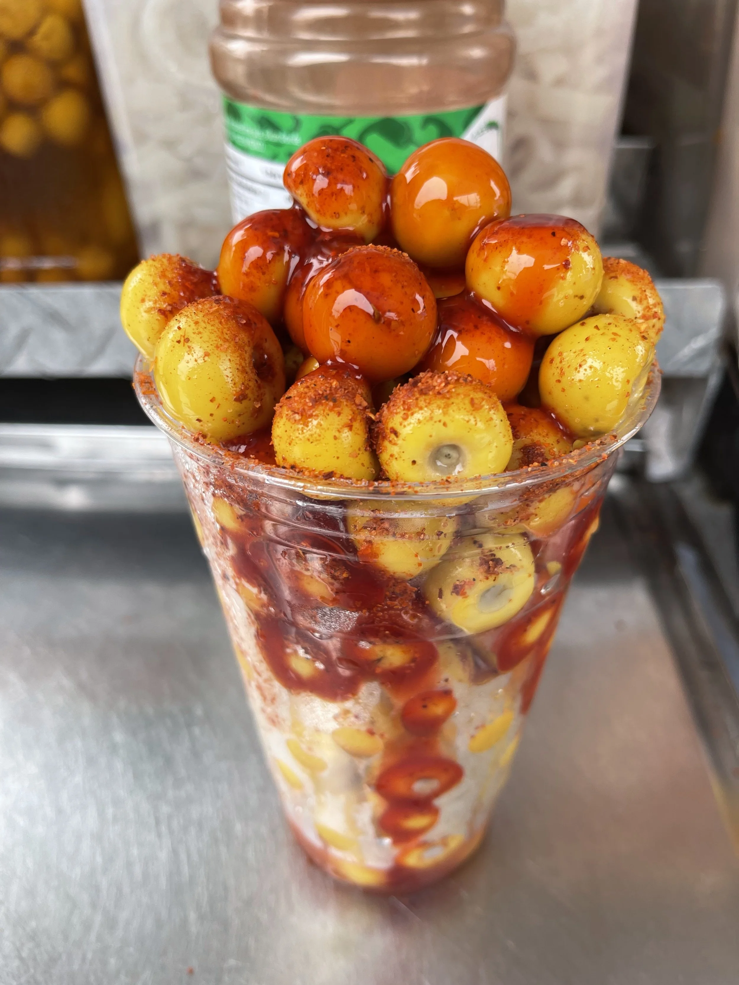 Close-up of a tall glass filled with yellow and orange grapes topped with a reddish-brown spicy seasoning and a drizzle of hot sauce.