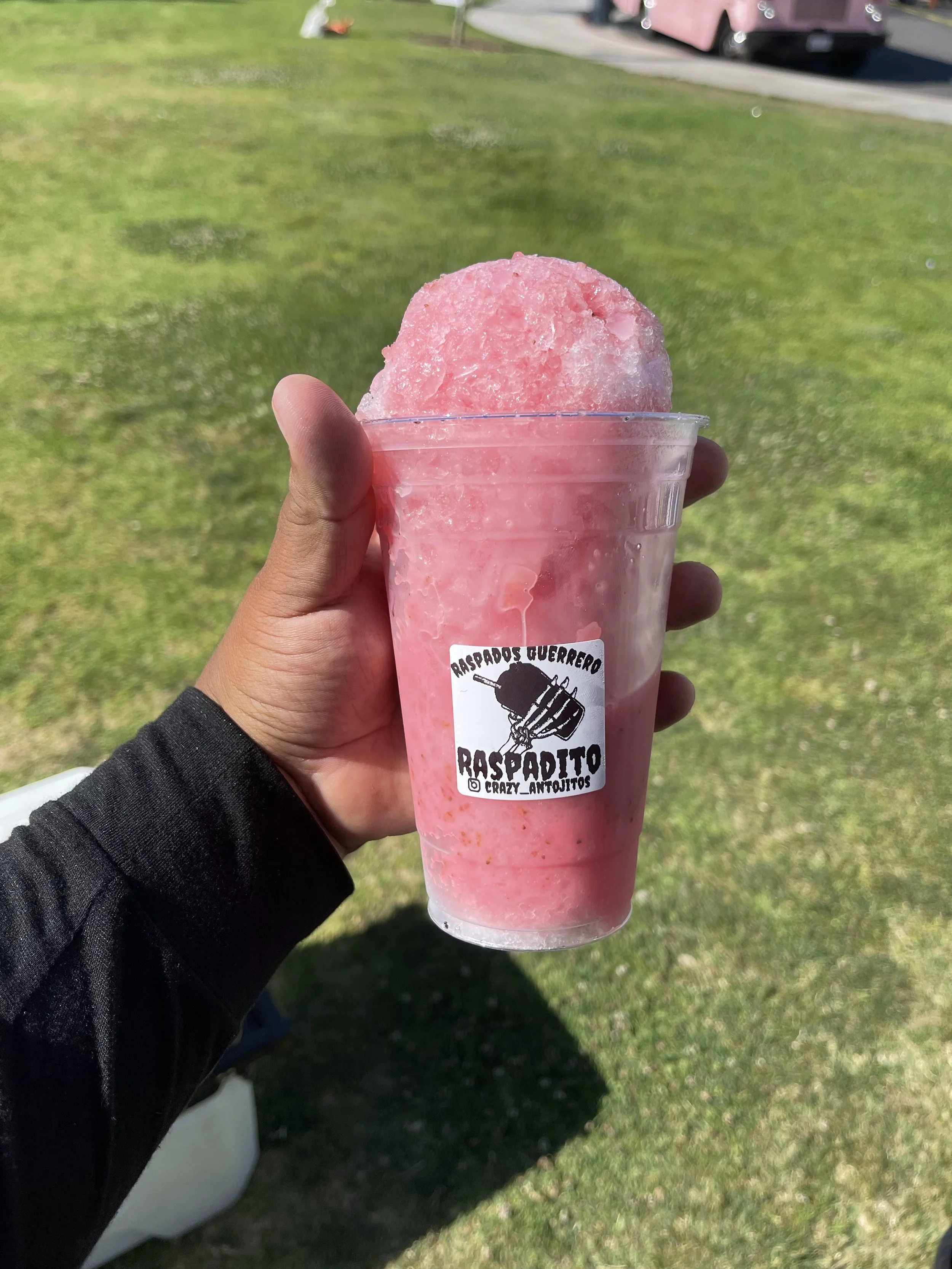 Hand holding a pink snow cone in a clear plastic cup outdoors on grass with a pink vehicle in the background.