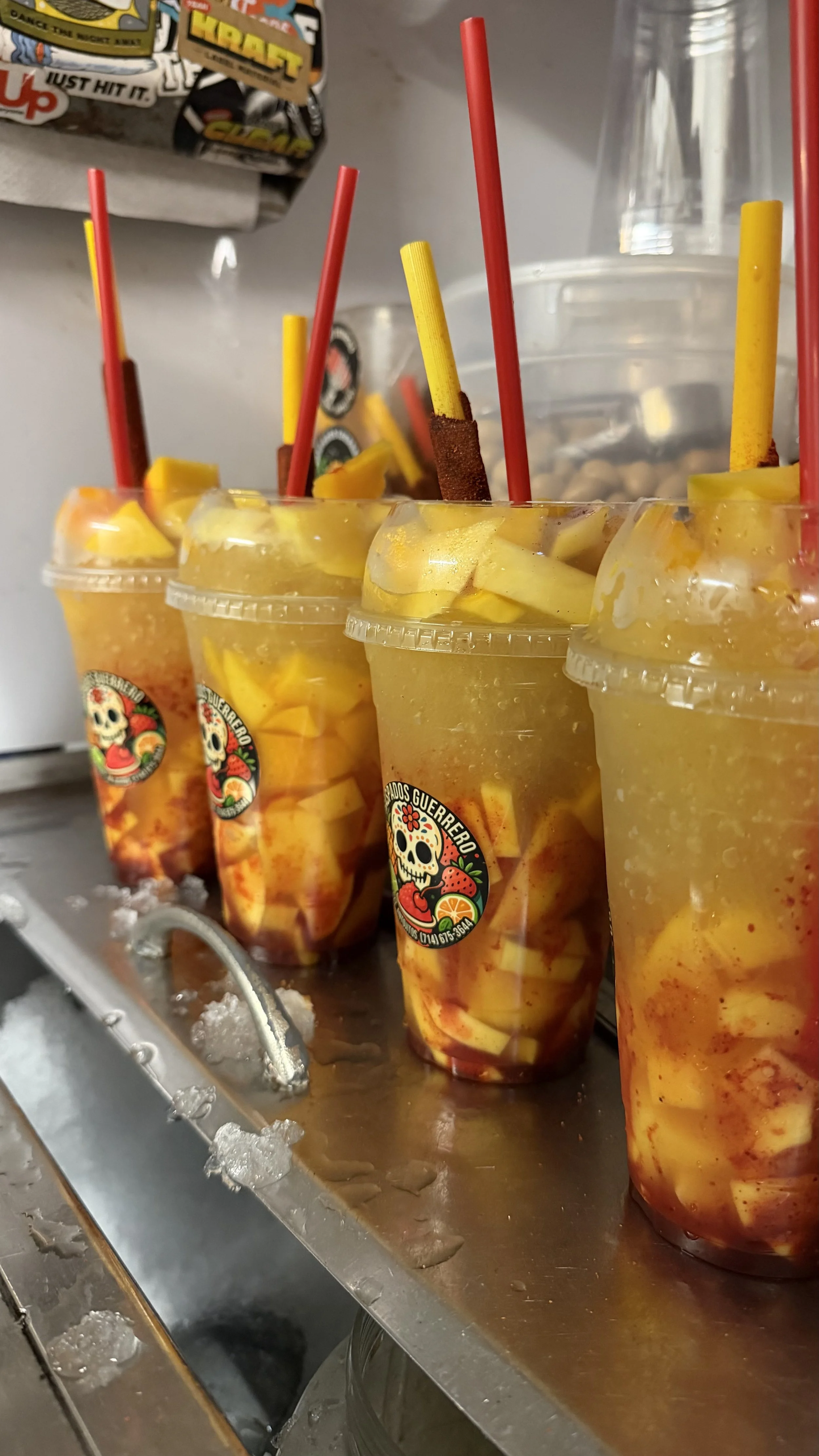 Four clear cups of fruit-based beverages with yellow and red straws, filled with chopped fruits like mango and strawberries, placed on a metal tray with ice, and a logo featuring a skull and colorful fruits.