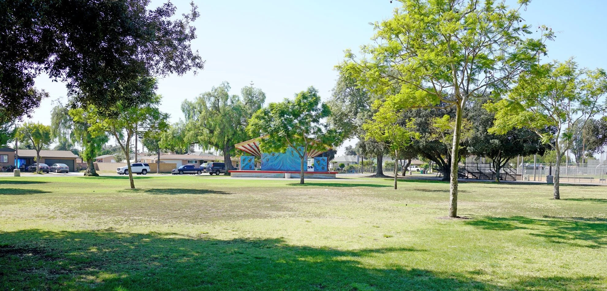 A spacious park with green grass, multiple trees, a colorful stage, and a playground area with steps, surrounded by a parking lot and residential houses.