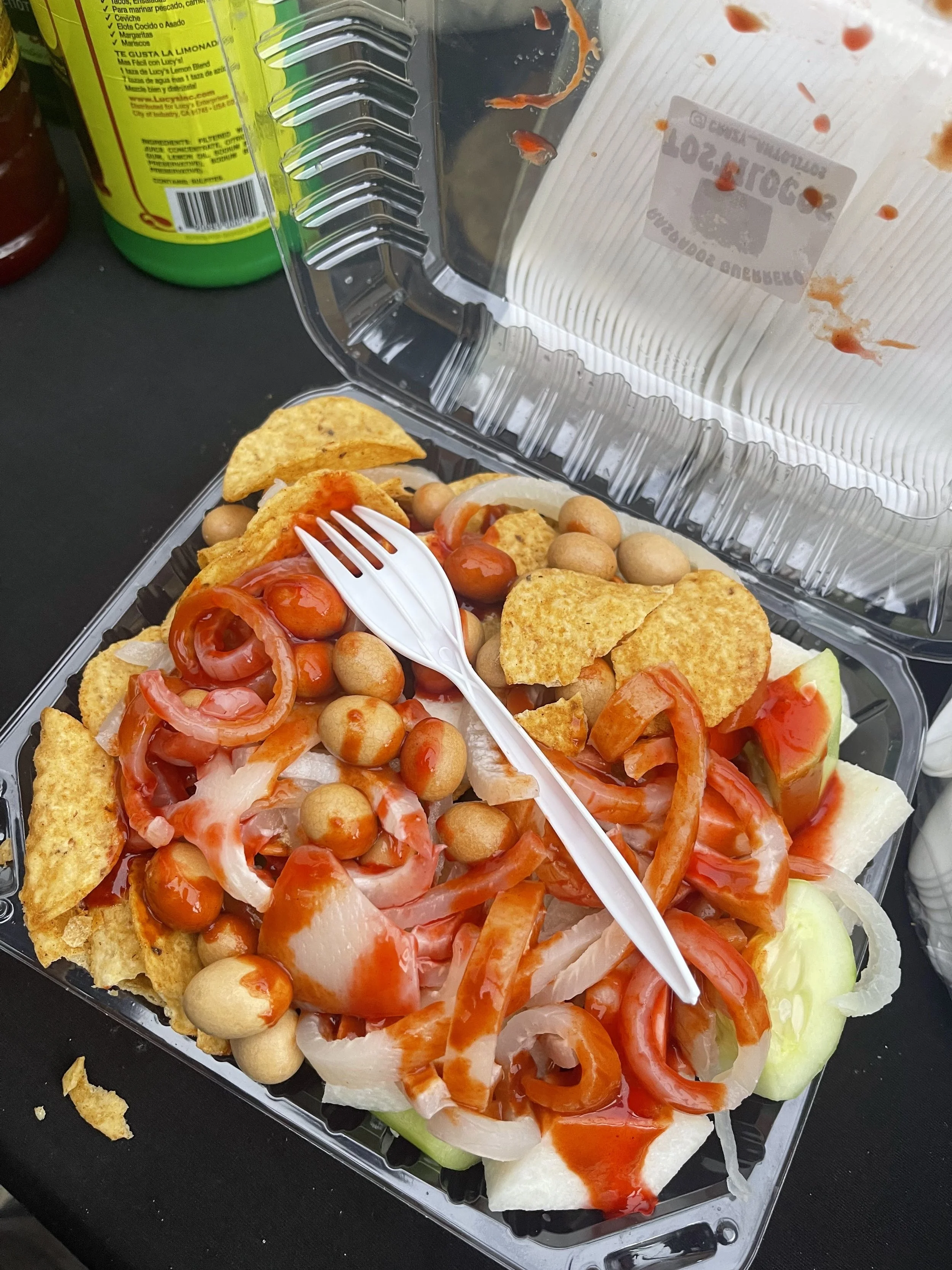 A plastic takeout container filled with a pizza topped with tomatoes, onions, chickpeas, and slices of cucumber, with some ketchup drizzled on top. The container is open, with a white plastic fork resting on the pizza. In the background, there is a yellow bottle of hot sauce and other condiments.