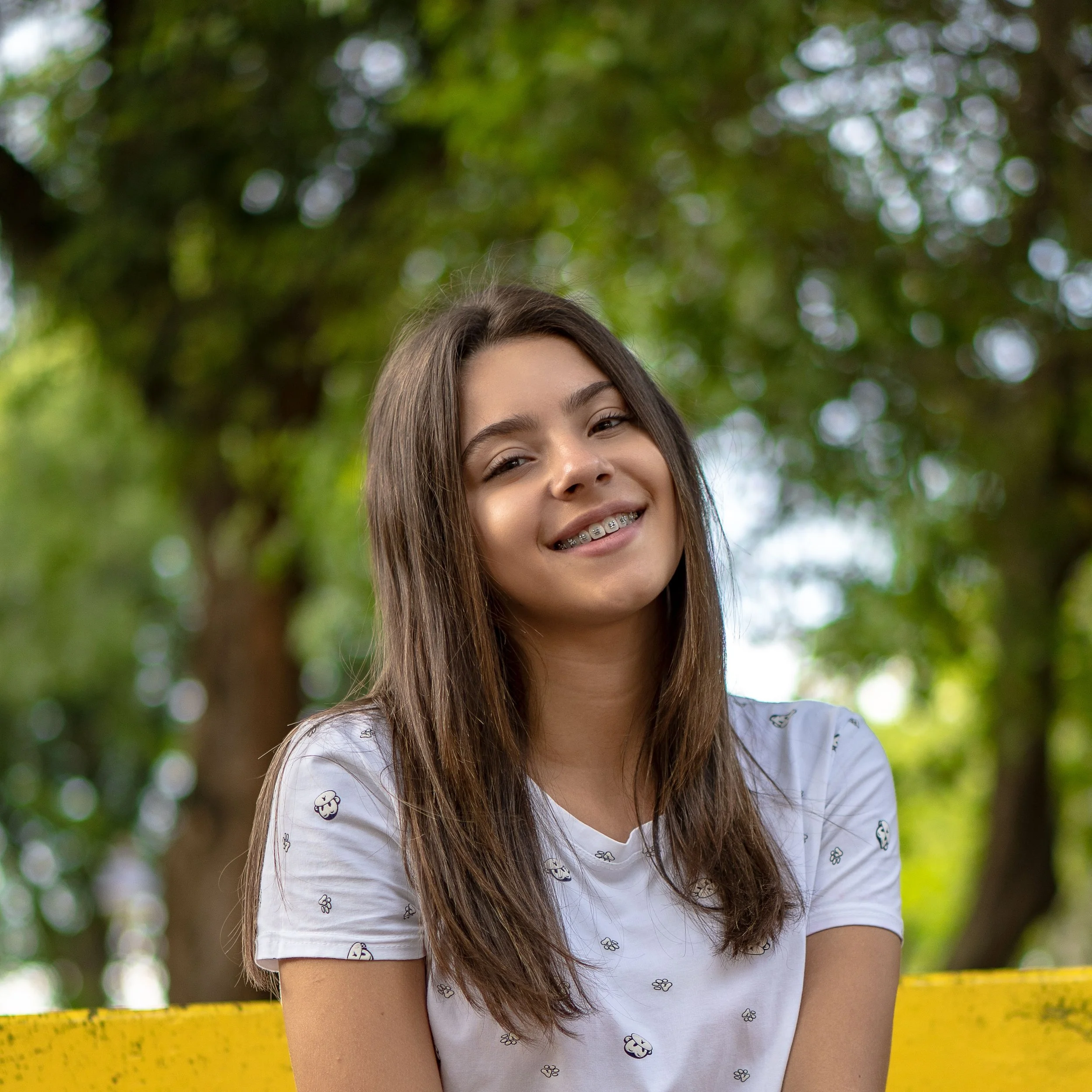 A young woman with long brown hair and braces smiling outdoors, with a blurred background of green trees and a yellow bench.
