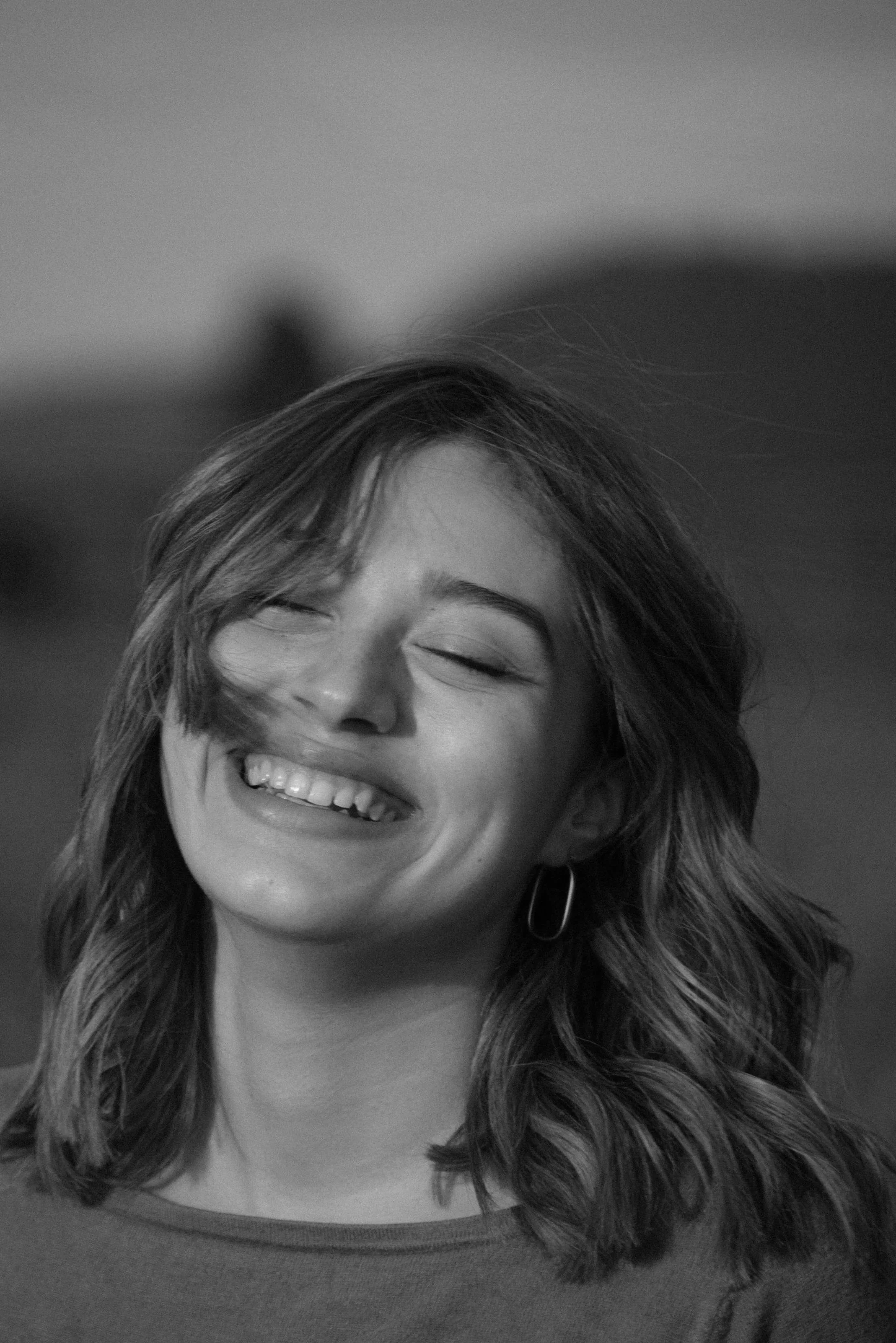 Black and white photo of a woman with shoulder-length wavy hair, smiling with closed eyes, wearing hoop earrings and a round-neck top.