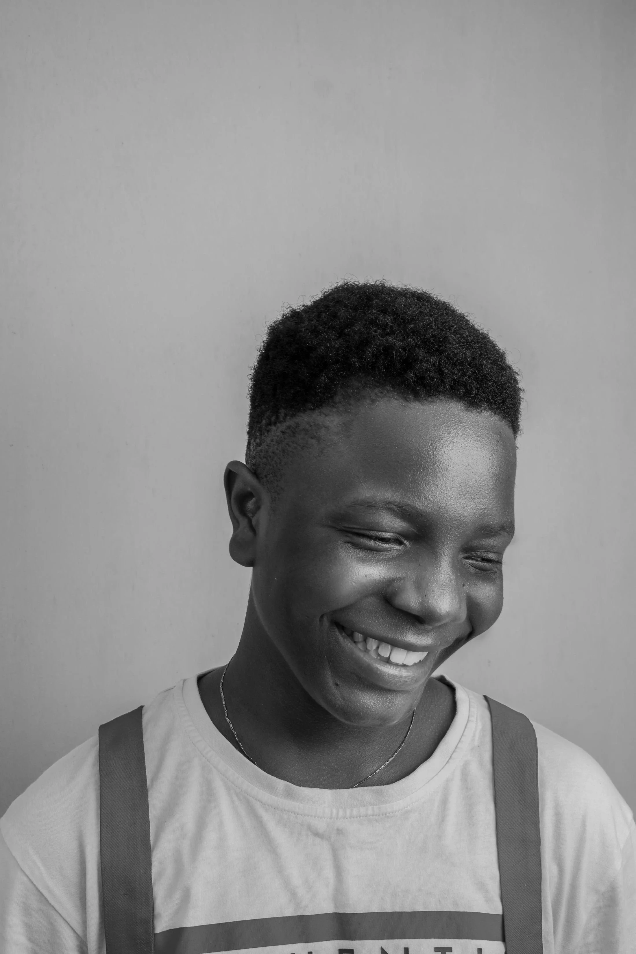 Black and white photo of a smiling person with short, curly hair, wearing a T-shirt and suspenders, with a plain background.