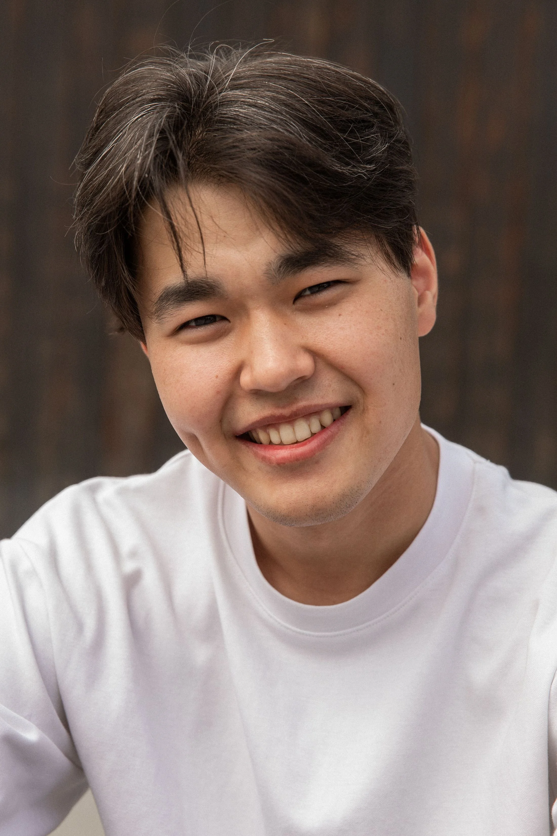 Close-up of a young man with dark brown hair smiling and wearing a white shirt.