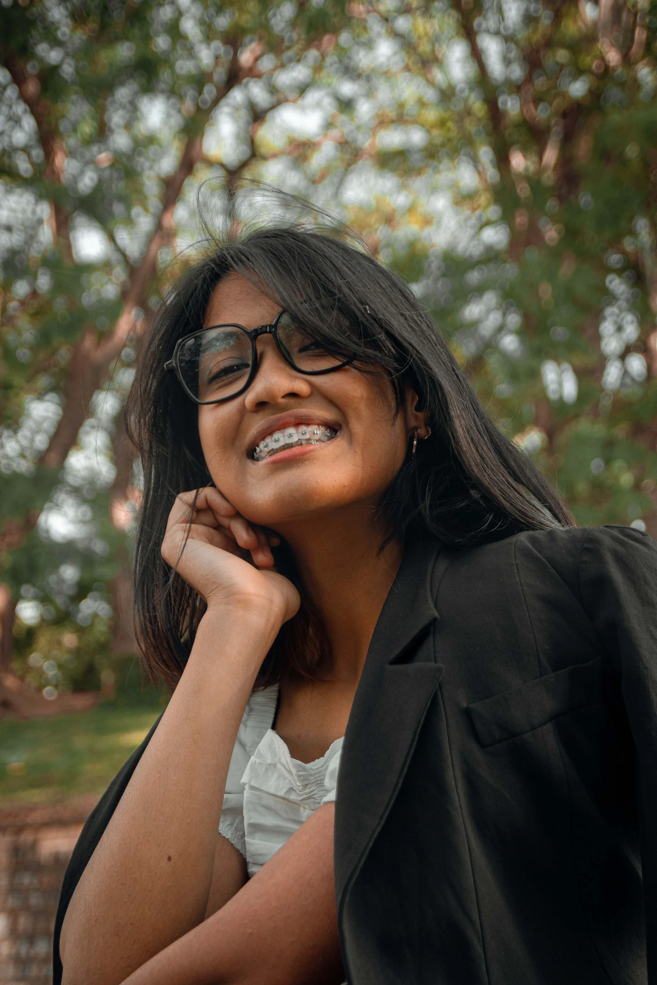 A woman with black hair and glasses smiling, resting her chin on her hand outdoors with trees and blurred background.