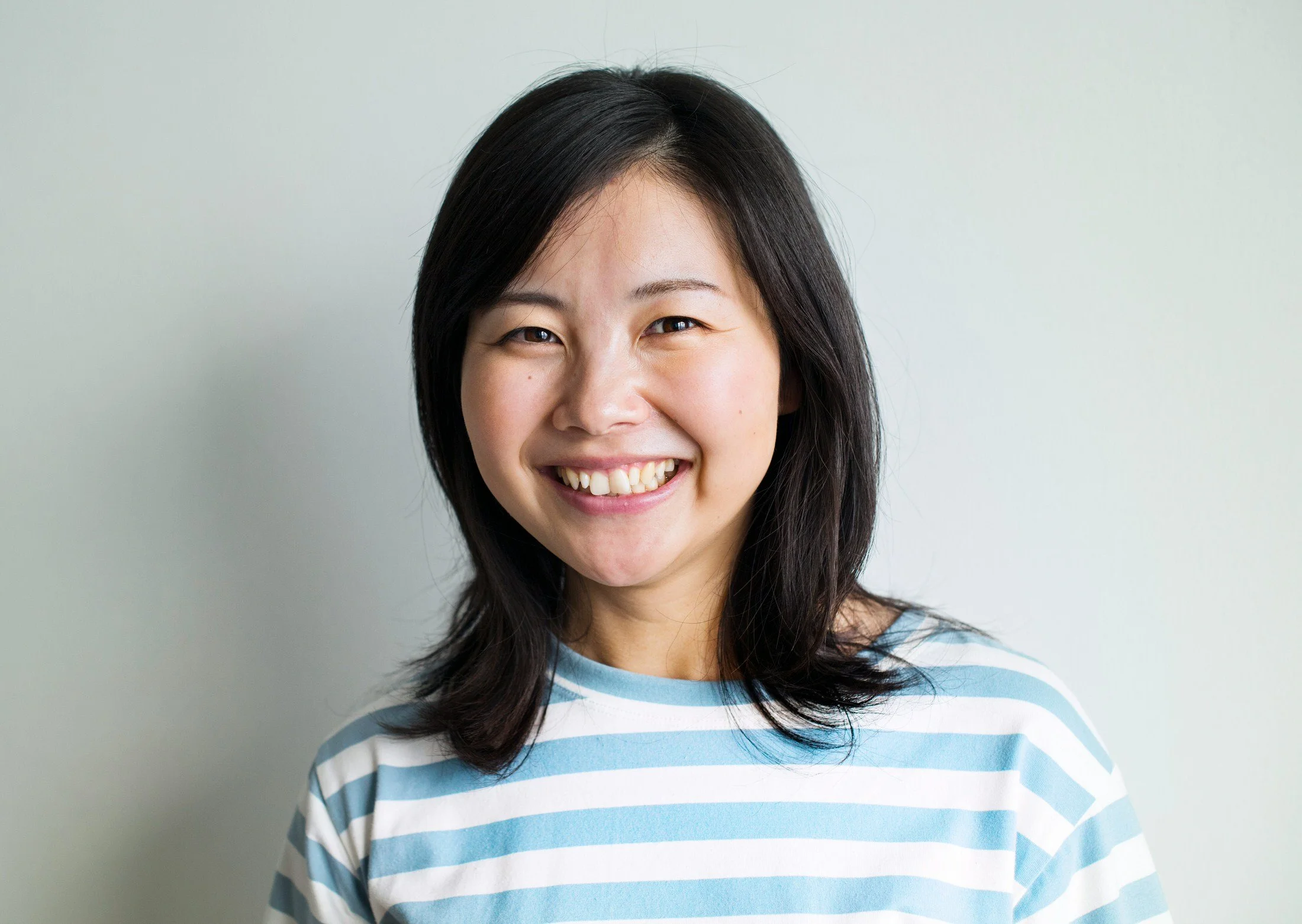 A woman with shoulder-length black hair smiling, wearing a blue and white striped shirt, standing against a plain light-colored background.
