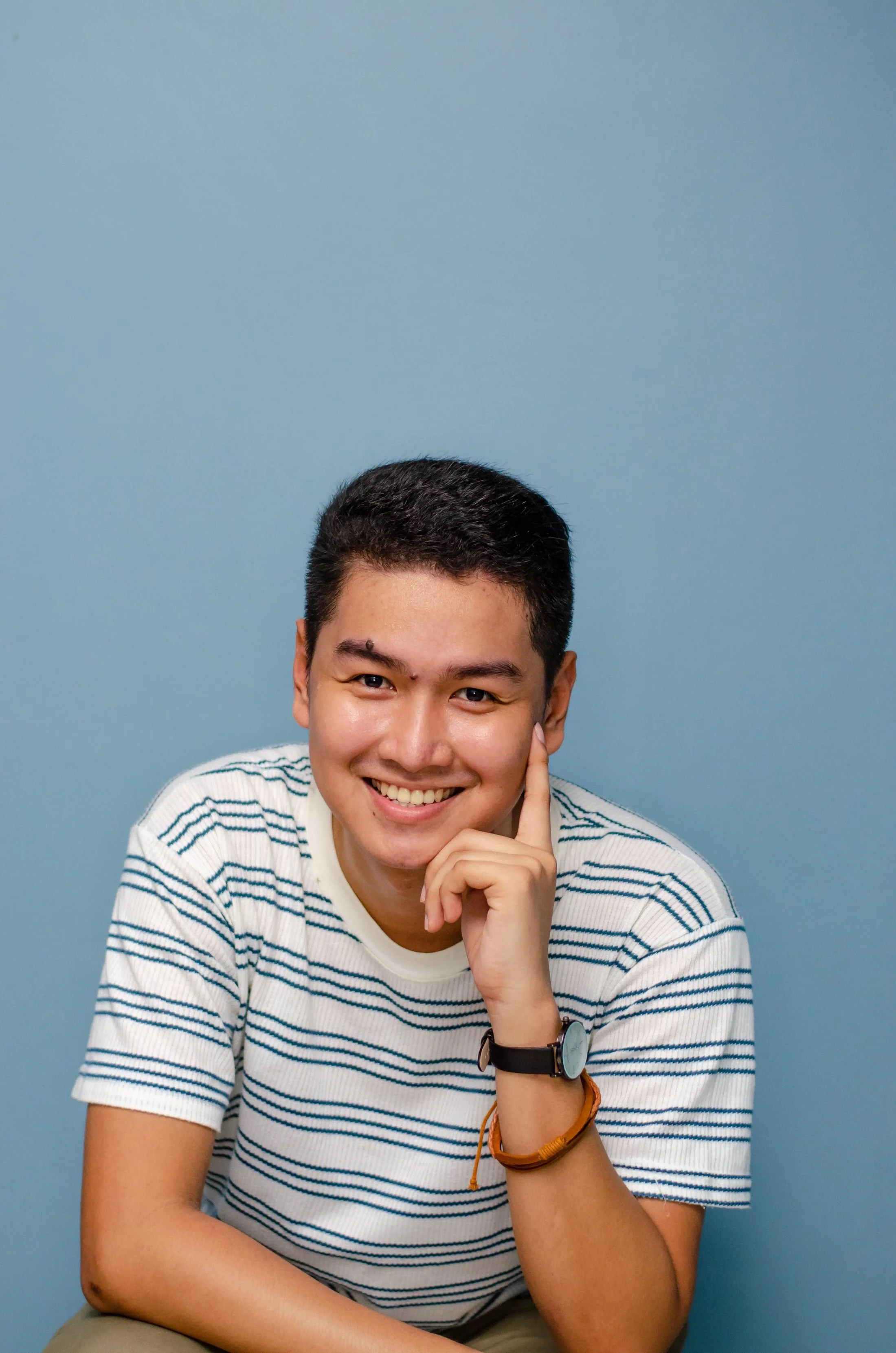 A young man smiling with his chin resting on his hand, wearing a striped t-shirt, a wristwatch, and bracelets, sitting in front of a blue background.