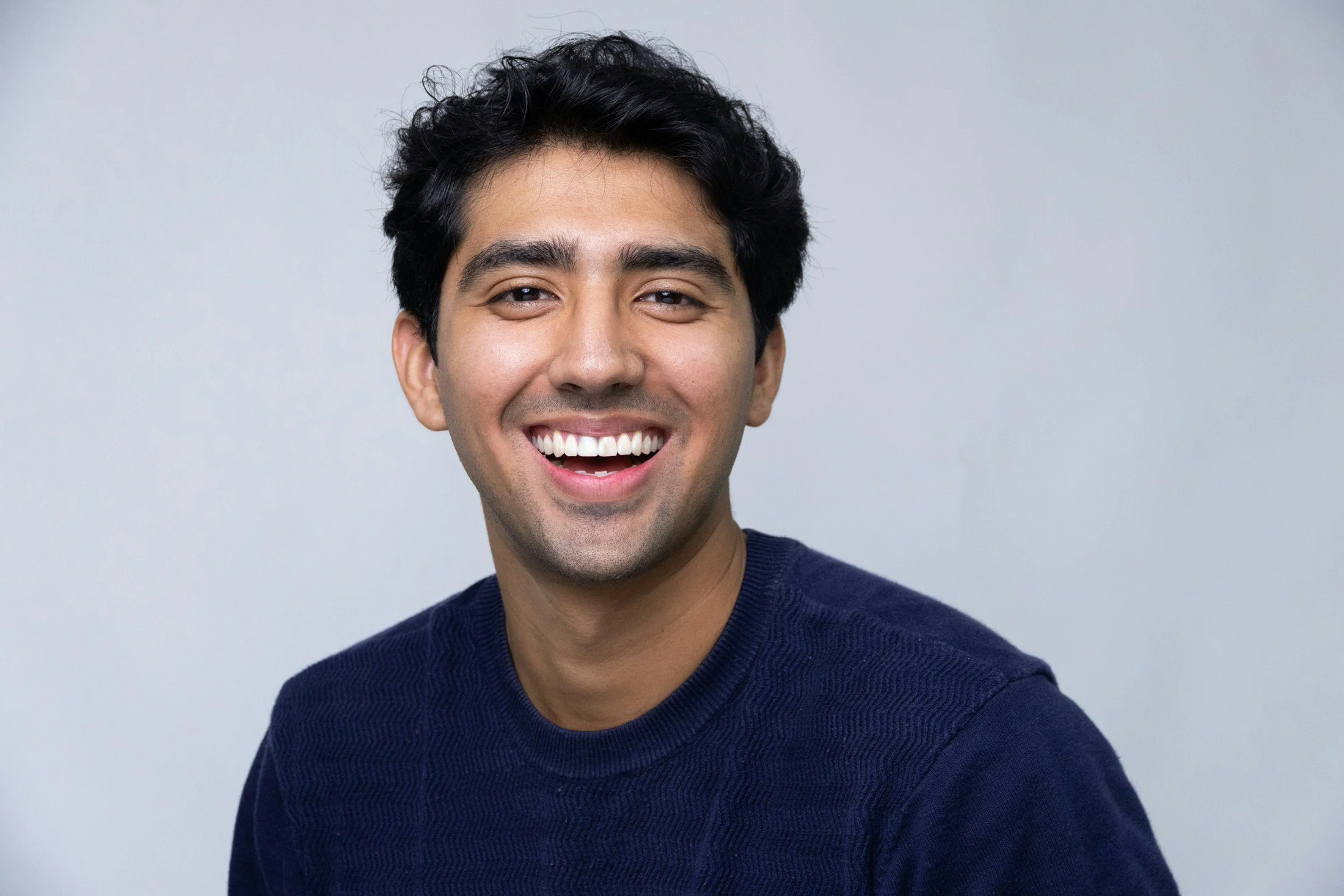 Close-up portrait of a smiling young man with dark hair wearing a navy blue sweater, against a light gray background.