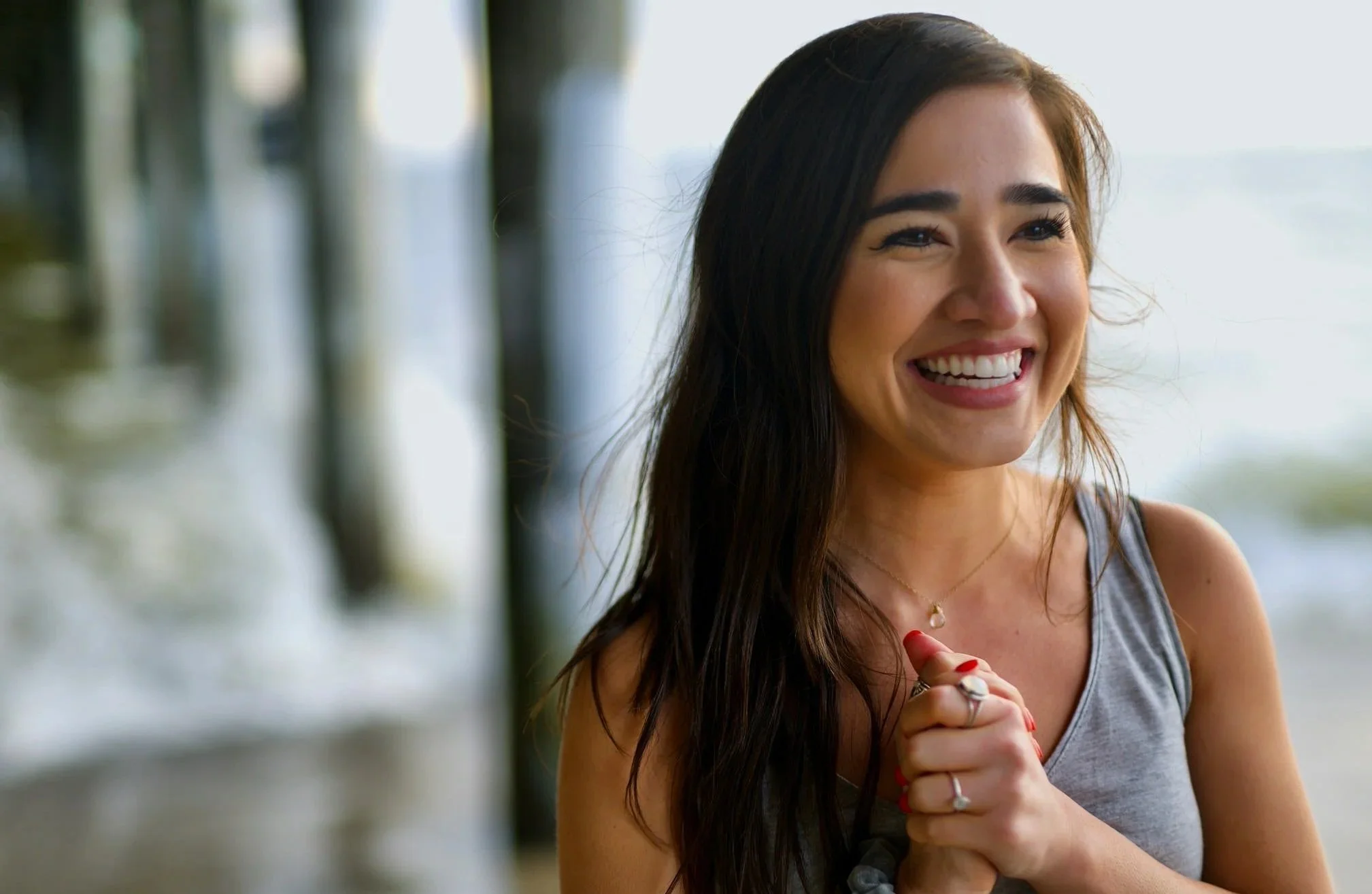 A woman with long dark hair smiling and clasping her hands together, wearing a gray tank top and jewelry, outdoors with blurred trees and sky in the background.