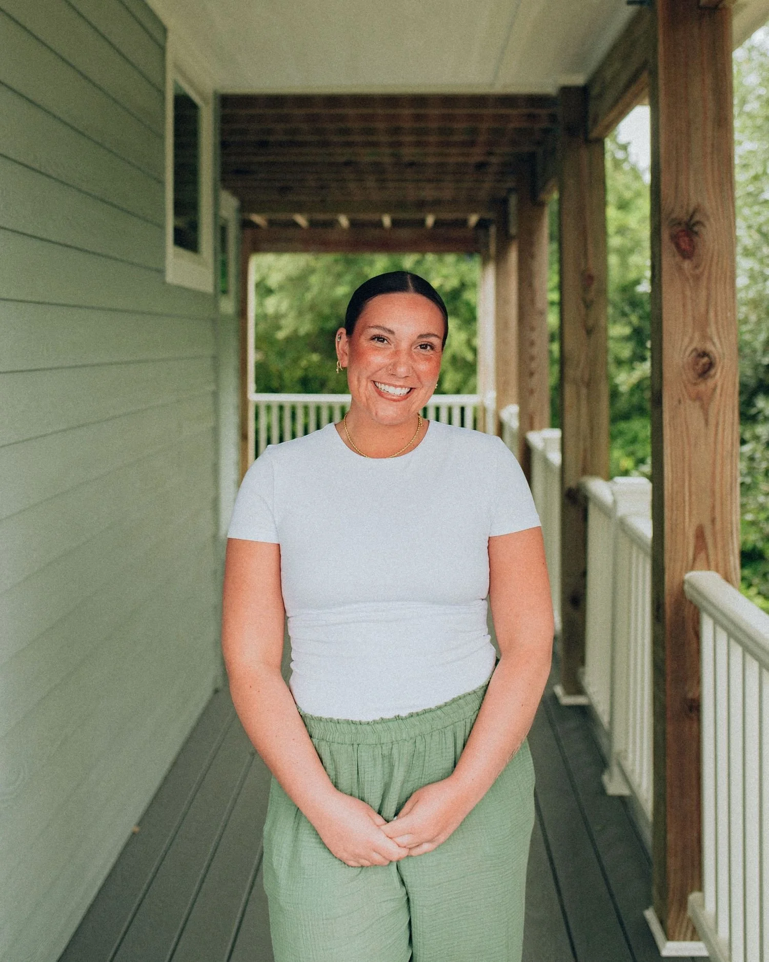 A woman smiling on a porch with green siding on one side, wooden beams above, and lush green trees in the background. She wears a white T-shirt and green pants.