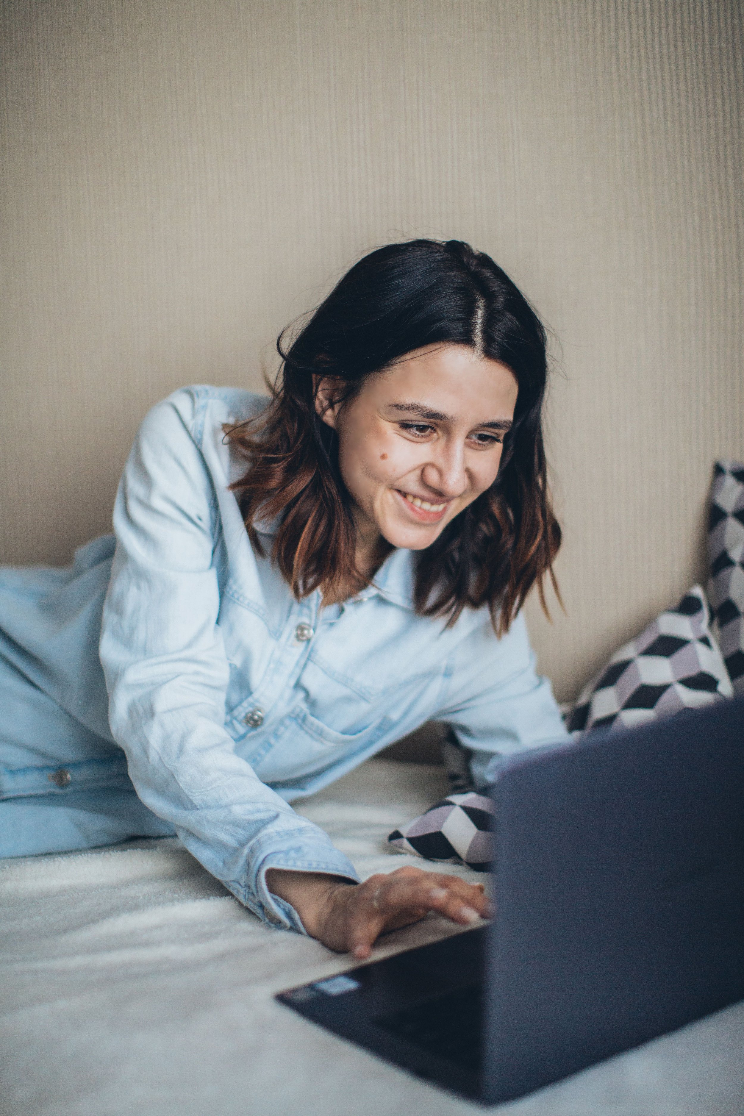 A young woman with dark hair and a smile, lying on her stomach on a bed, using a laptop.