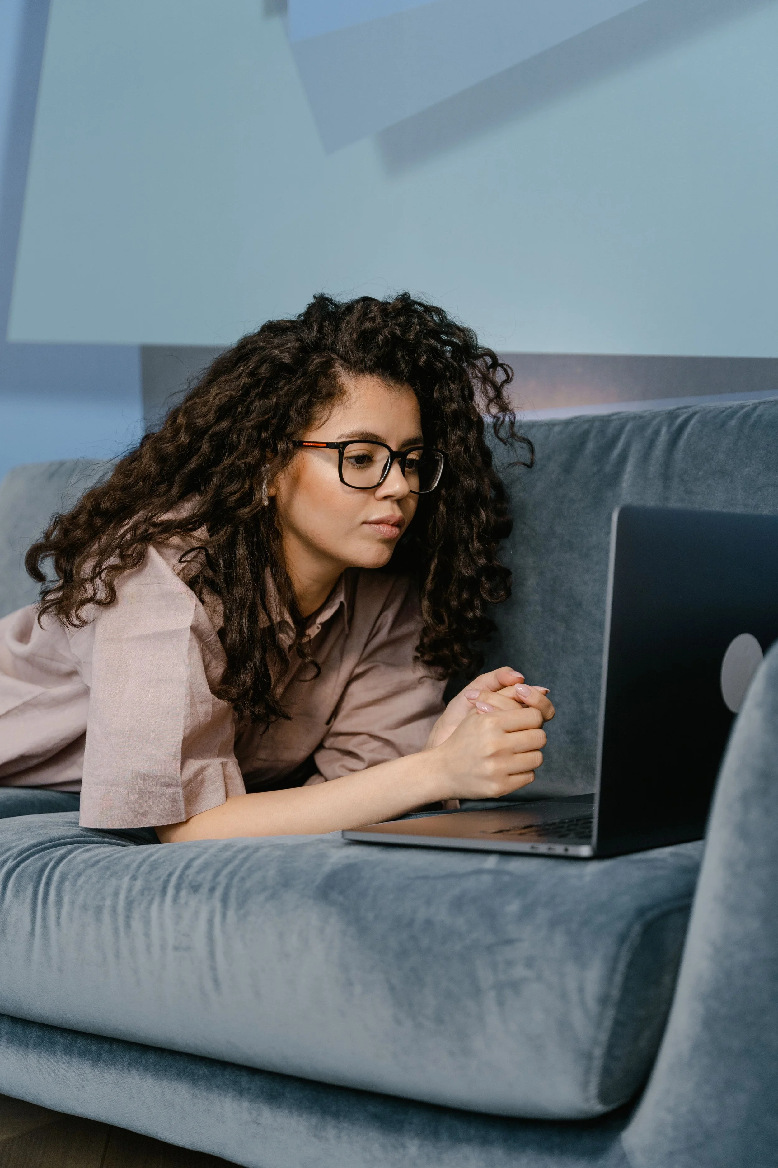 A young woman with curly hair and glasses is sitting on a gray couch, working on a laptop.