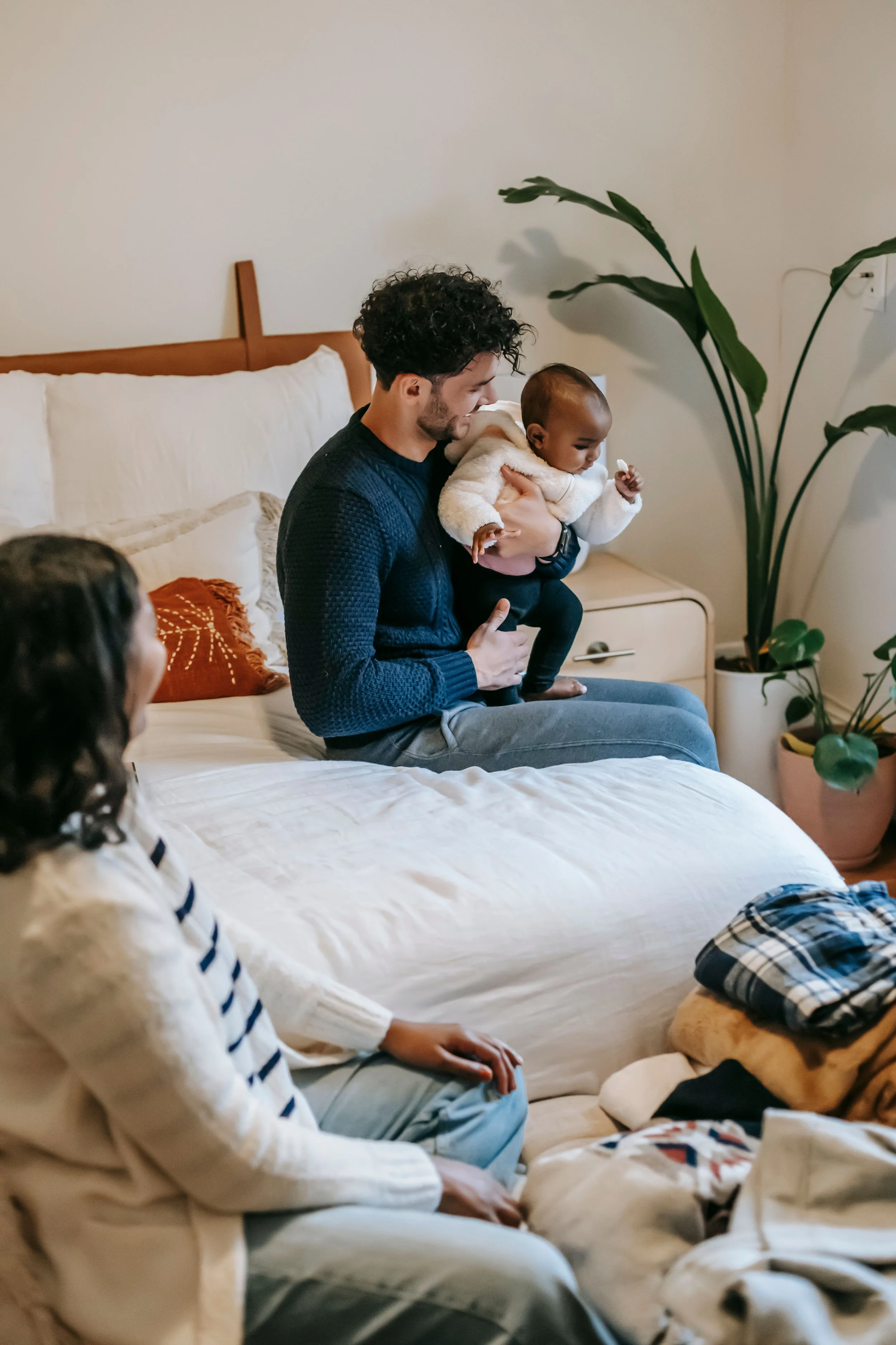 A man holding a baby and smiling in a bedroom with a bed, pillows, a large plant, and a child sitting on the bed.