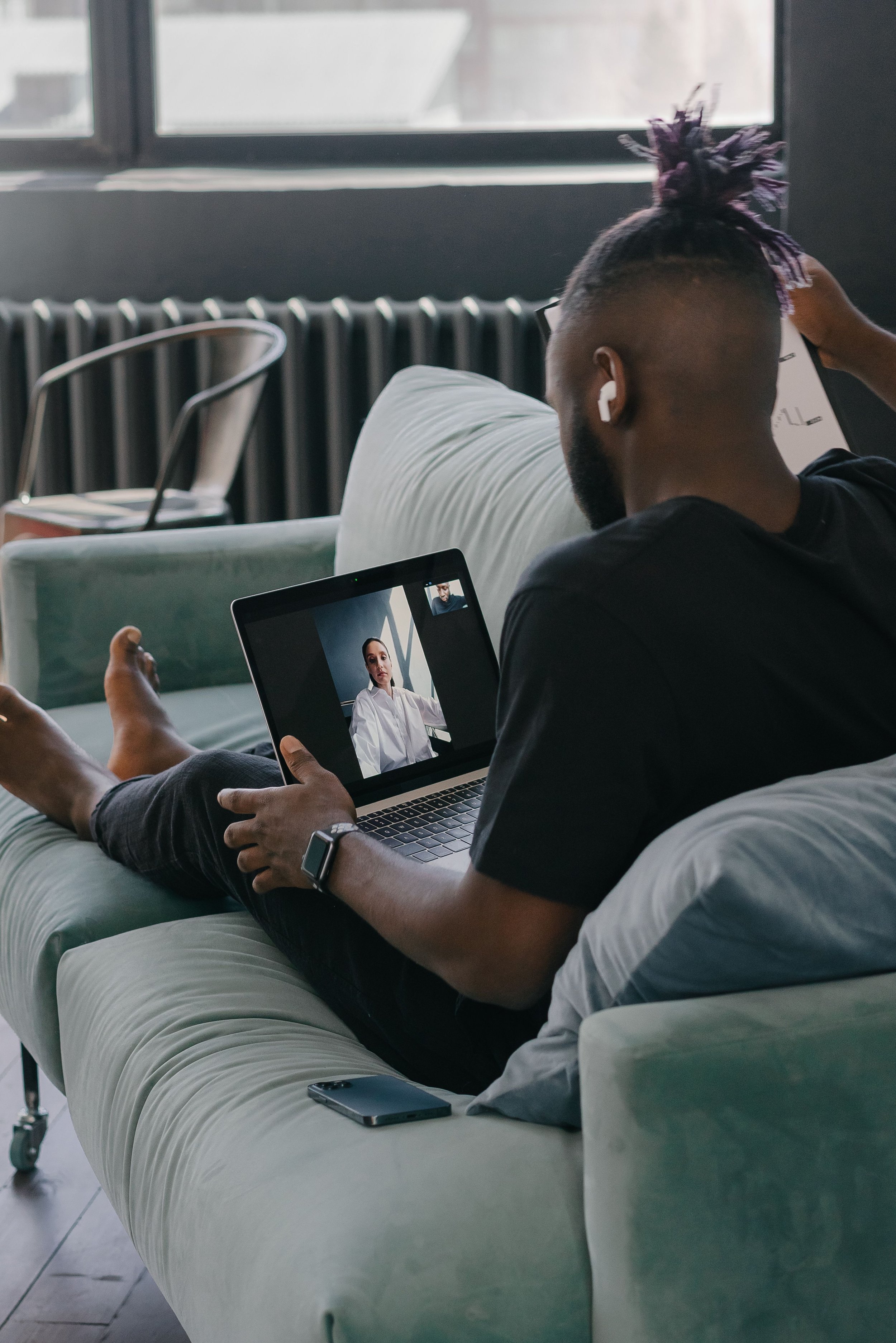 A man with purple hair styled in a topknot, wearing a black shirt and white wireless earbuds, is sitting on a light green couch, holding a laptop showing a video call with a woman in a white shirt. There are chairs and a radiator near a window in the background.