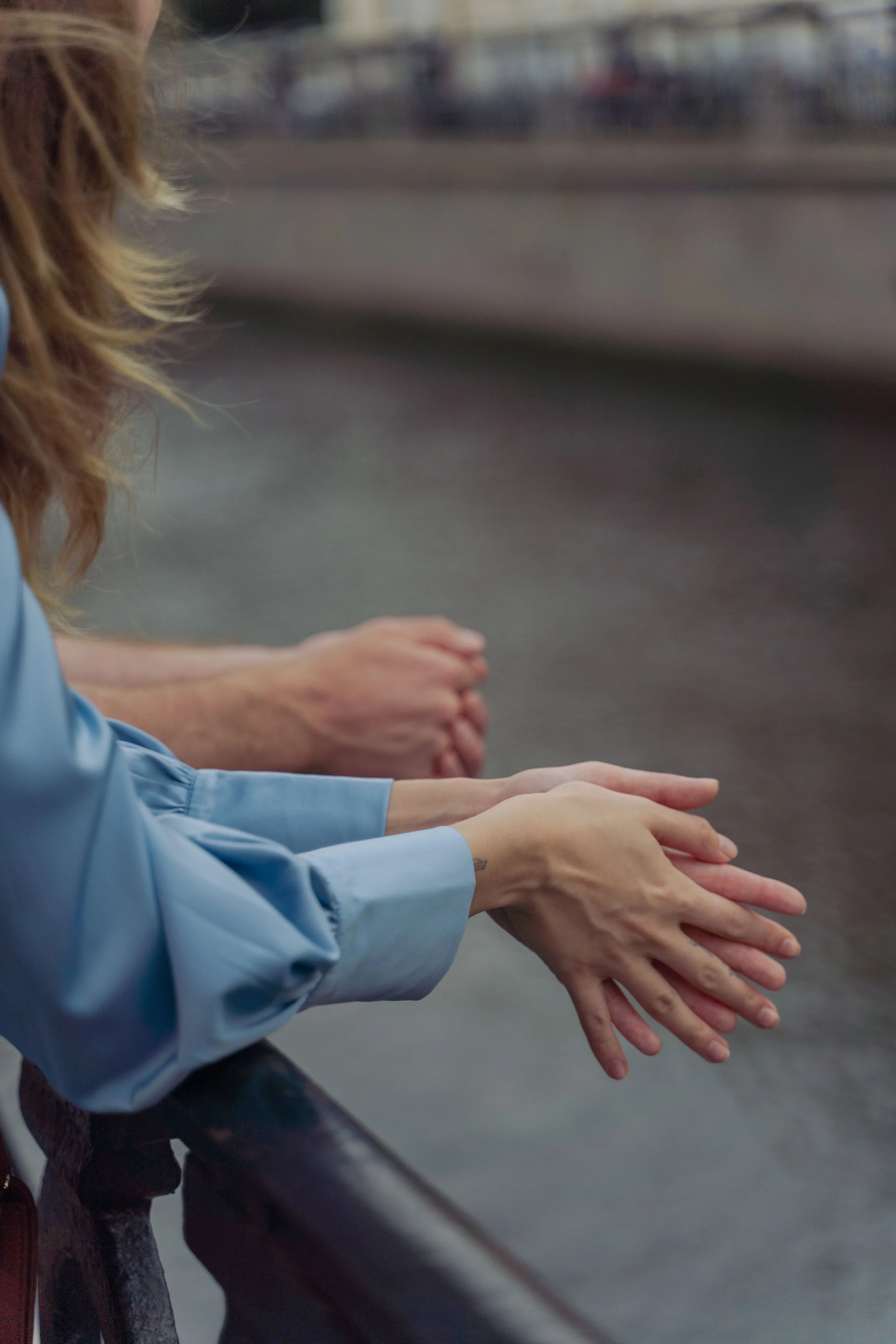 Multiple pairs of hands are placed on a metal railing in a row. One person is wearing a light blue shirt, and another hand is visible at the far end of the row. The background is blurred, indicating an outdoor setting.