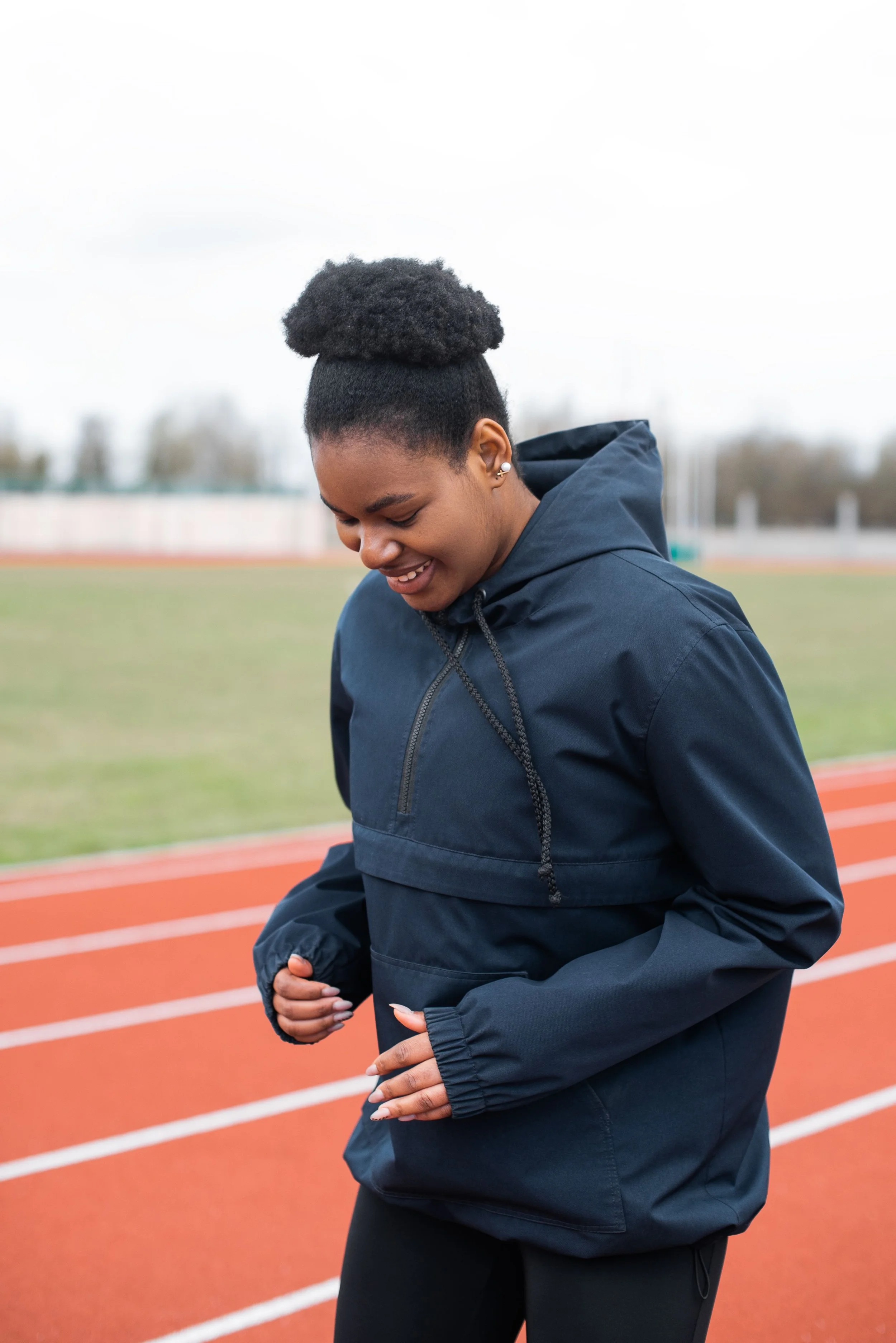 A young woman in a navy blue athletic tracksuit on a running track with her head bowed and eyes closed, smiling softly, with her hands resting on her stomach.