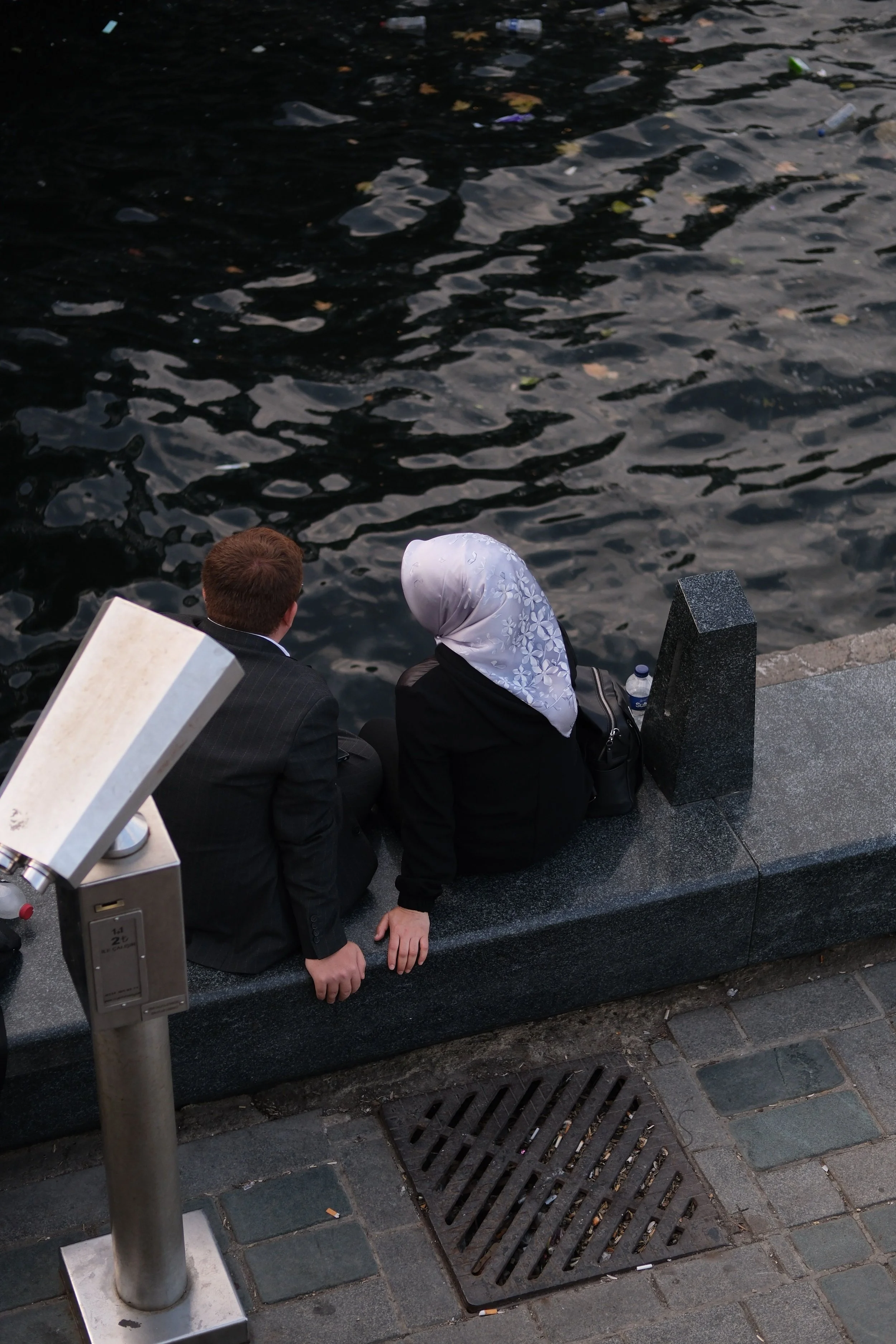 Two people are sitting on a stone ledge near water, seen from behind, with a parking meter in the foreground.
