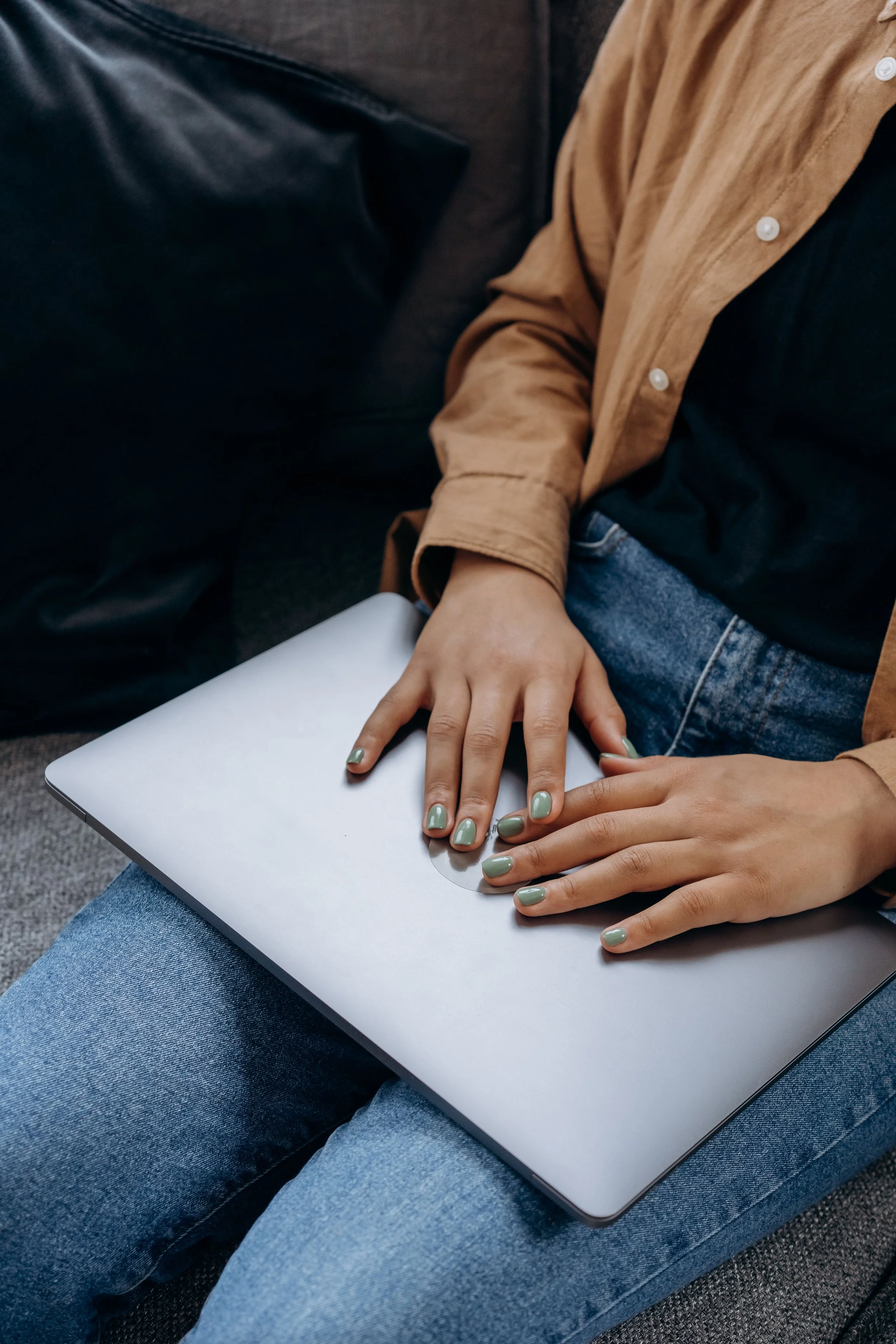 Person sitting on a gray couch, wearing blue jeans and a black A-line shirt, with a laptop resting on their lap. Their right hand has green nail polish and is holding the laptop, while their left hand is resting on top of the laptop.
