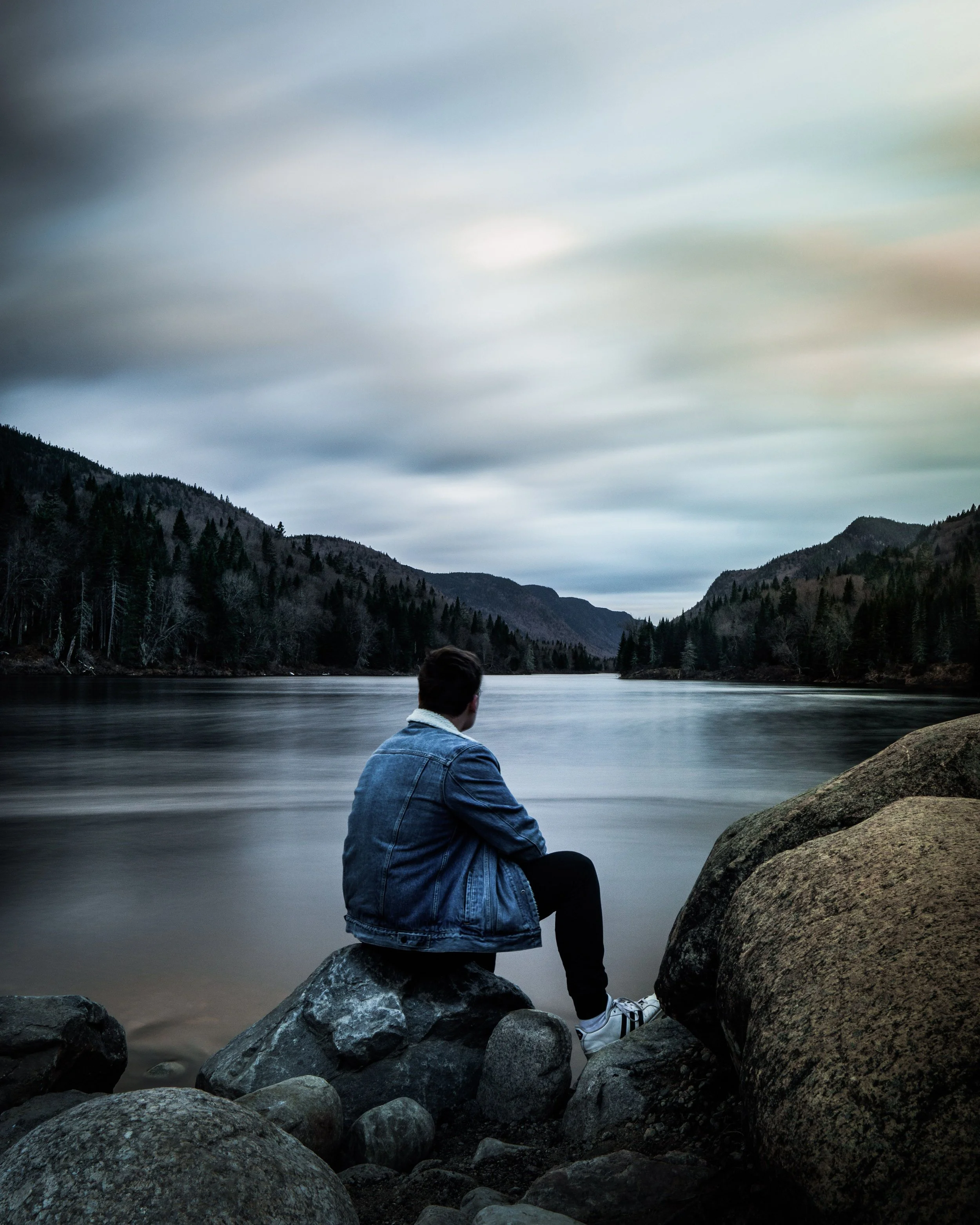 Person in denim jacket sitting on rocks by a calm lake with mountains and overcast sky in the background.