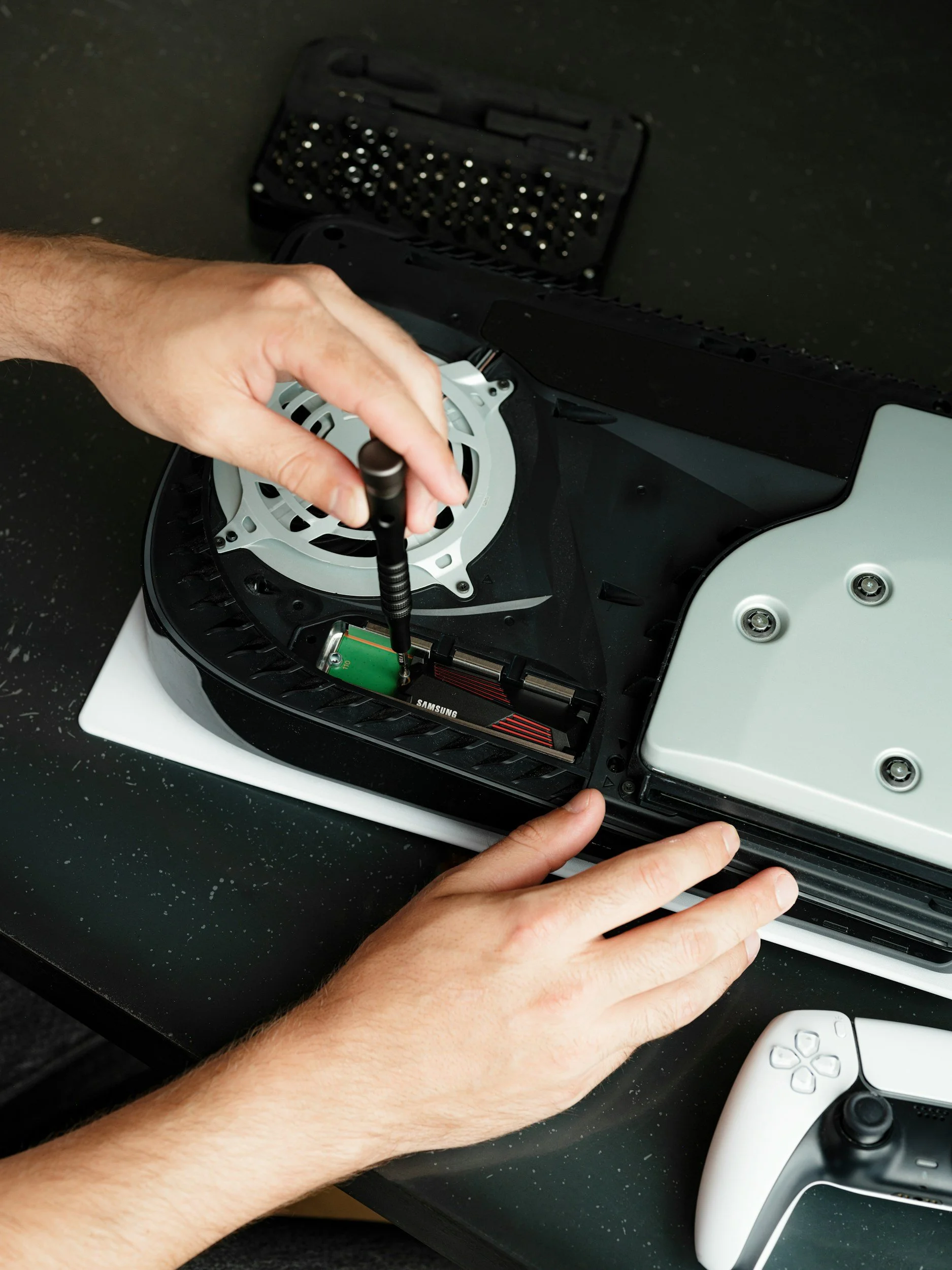 Person's hands repairing or assembling a gaming console with a screwdriver, with a white gaming controller nearby.