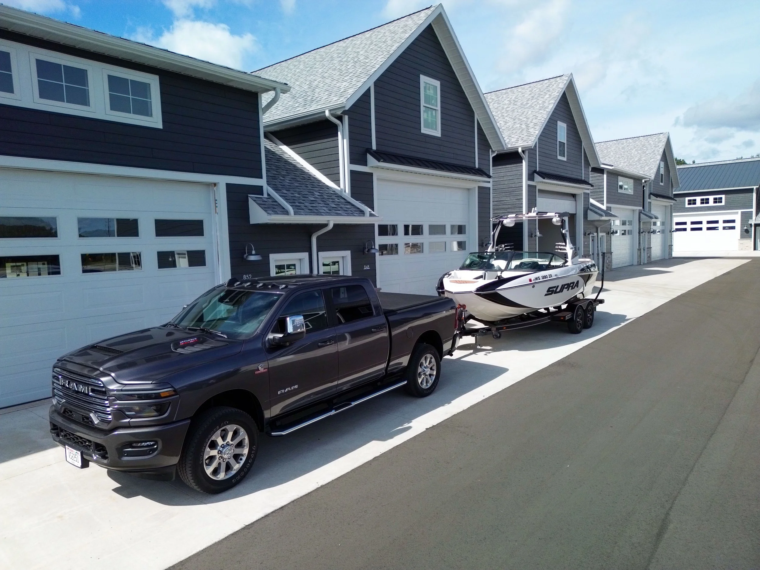 A modern residential driveway with a dark gray pickup truck towing a boat, with multiple attached garages and a row of townhouse-style homes with gabled roofs in the background.