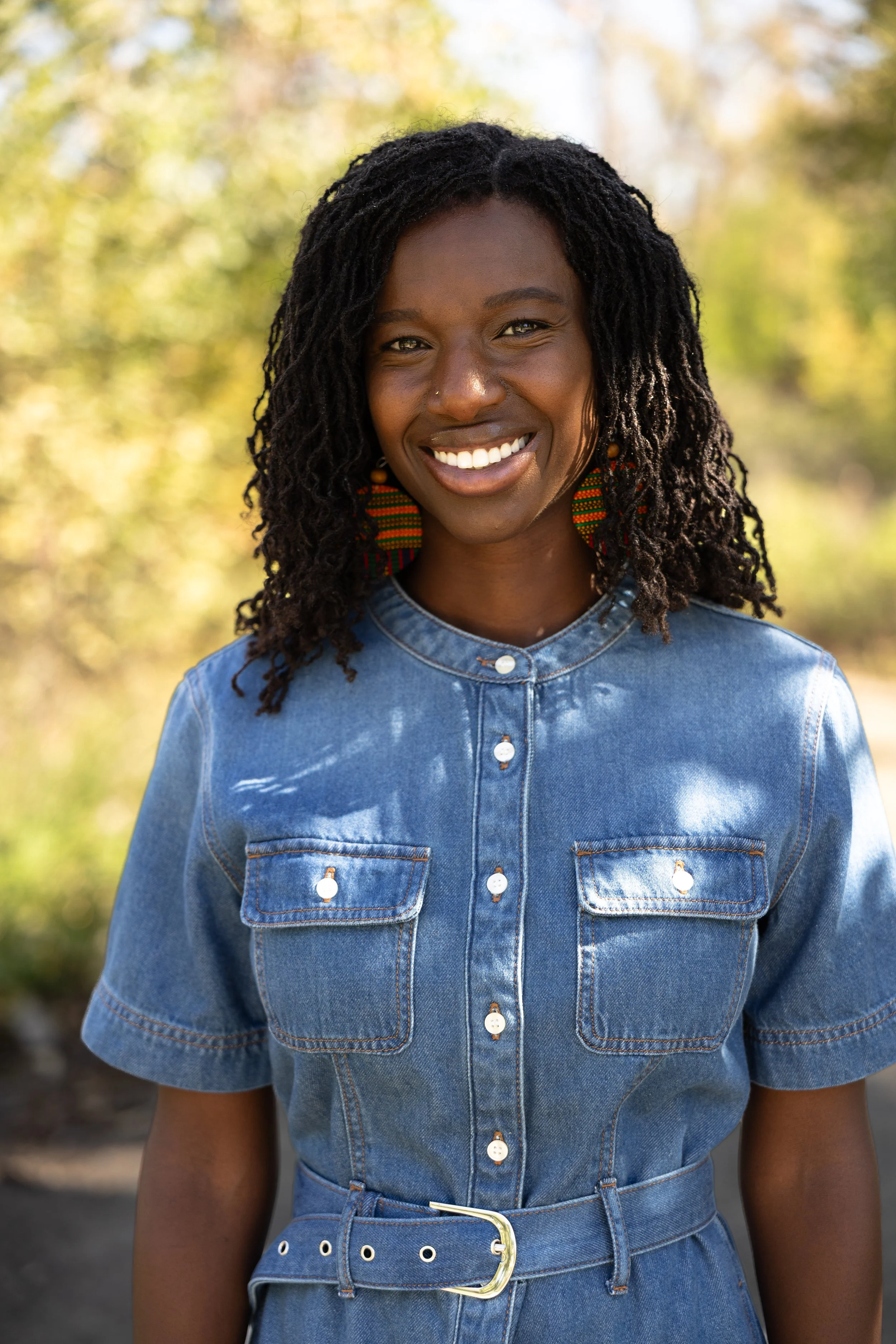 Portrait of a smiling Black woman with dreadlocks, wearing a denim shirt and a matching denim belt, standing outdoors with blurred autumn trees in the background.