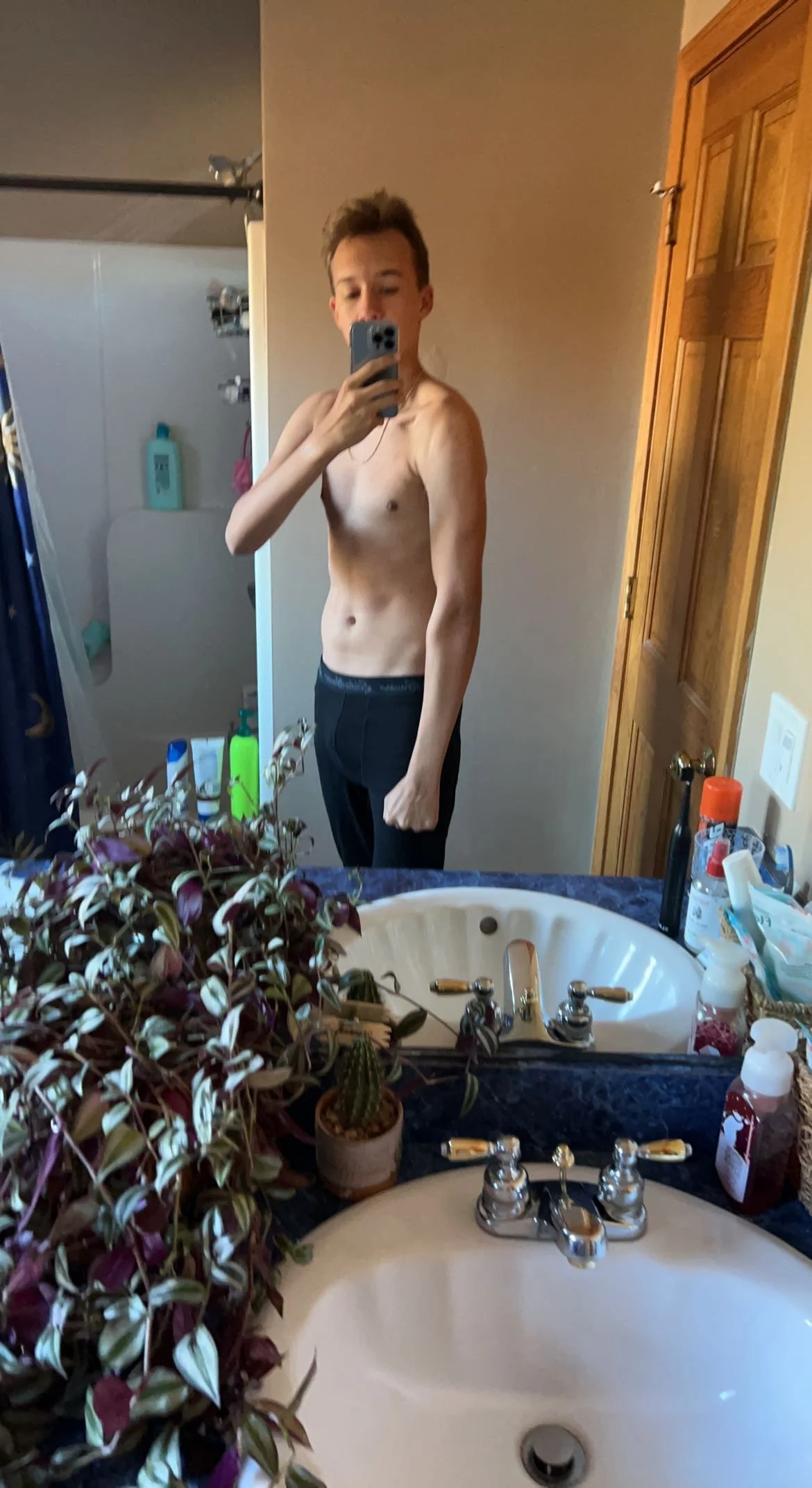 A shirtless young man taking a selfie in a bathroom mirror. The bathroom has a sink with various toiletries on the counter and a towel on a hook in the background.