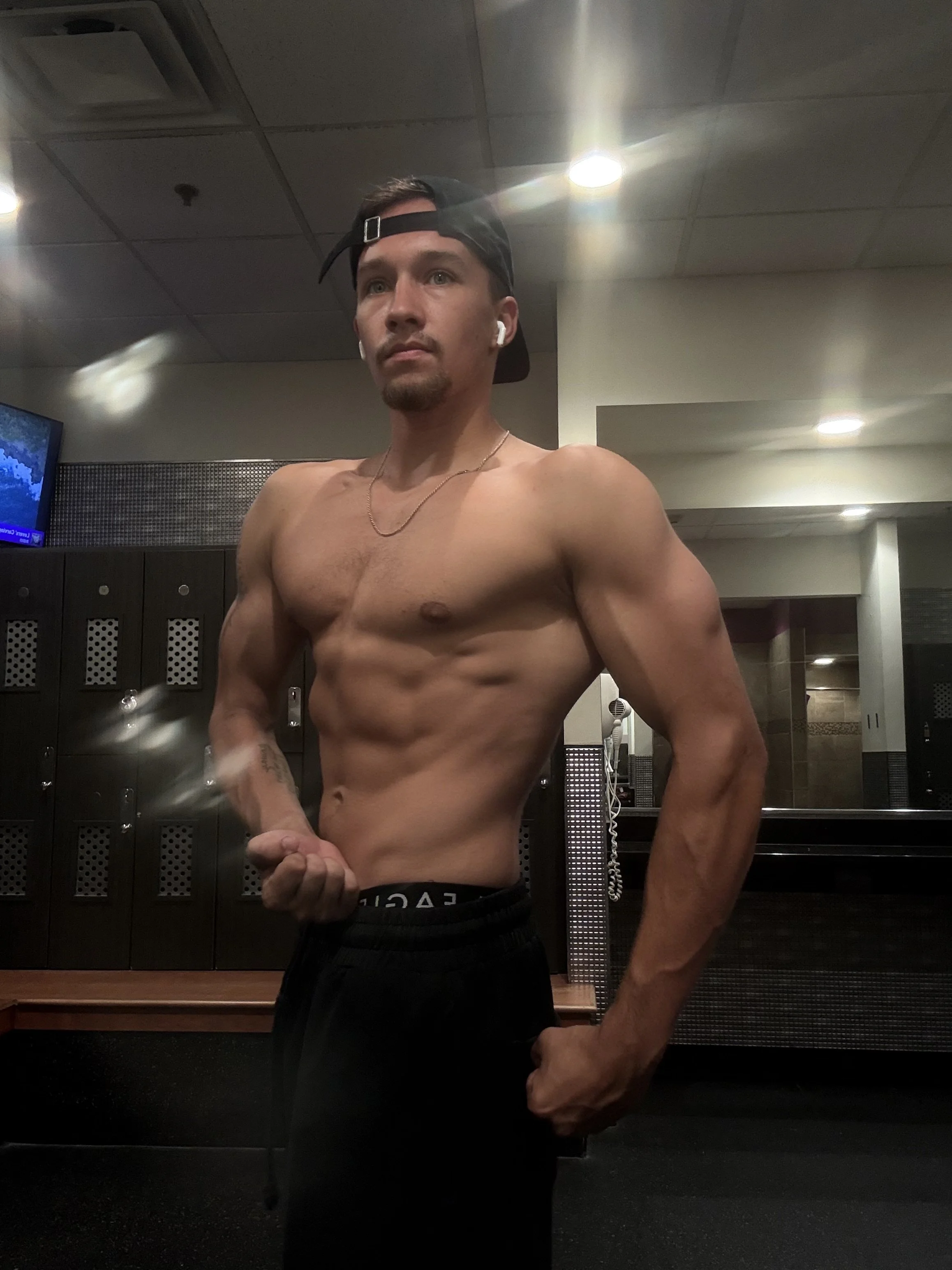 A shirtless young man with a beard and mustache taking a mirror selfie in a gym locker room. He is wearing a backward black cap, wireless earbuds, a thin chain necklace, and black shorts.