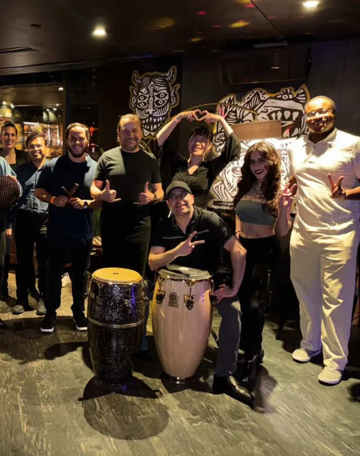 A salsa class in a restaurant or club, with two salsa instructors in front, making peace signs and a heart shape with their hands. Colorful artwork on the wall, with some smiling and enjoying the scene. A pair of congas is visible.