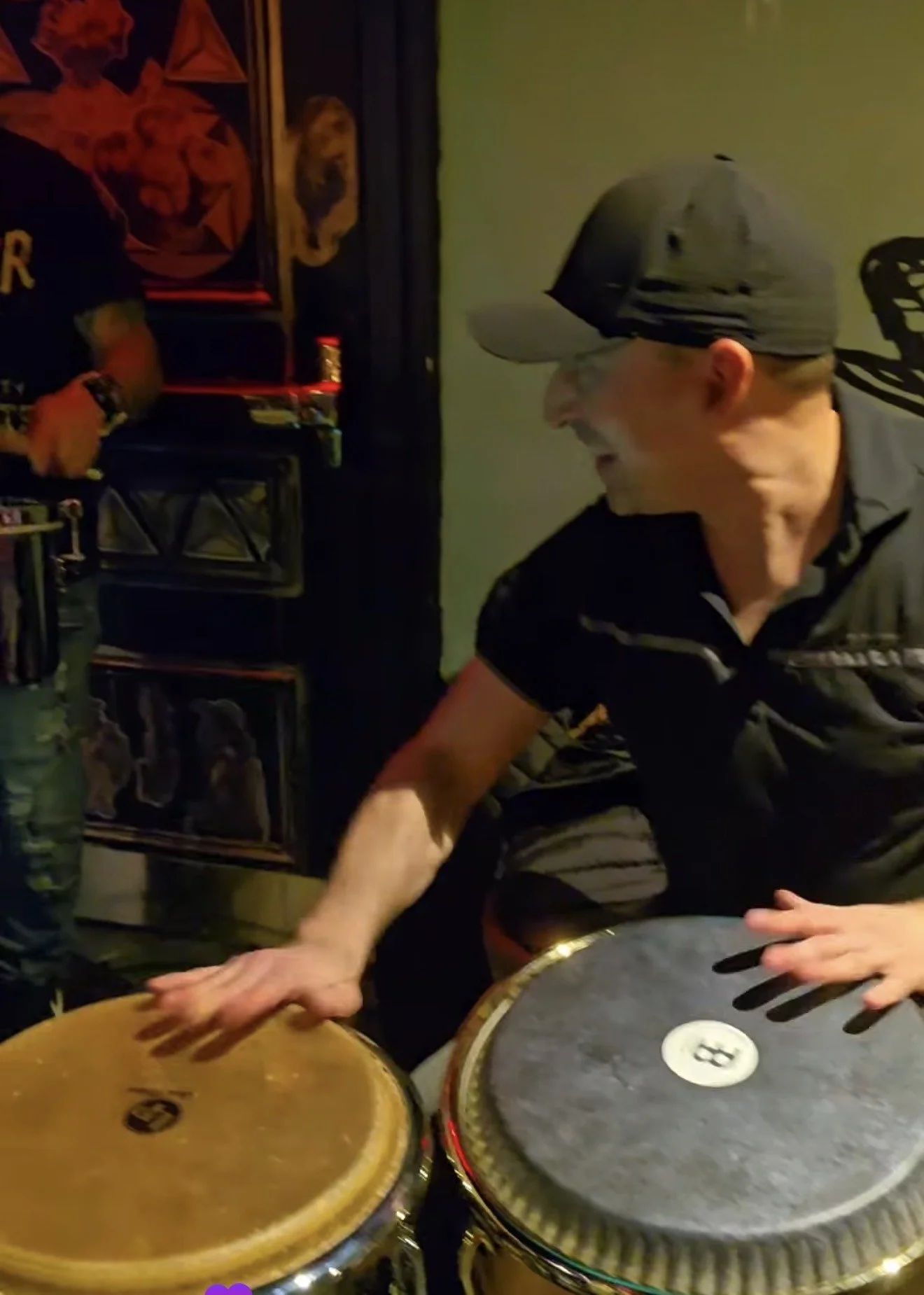 A person wearing a cap and glasses playing conga drums, with another person standing nearby in a dimly lit room that has shelves with records or books and framed artwork on the wall.