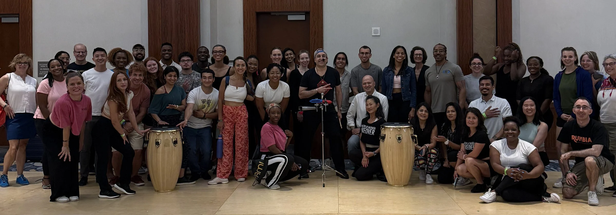 Group of diverse people from a salsa class posing together in a large room with some musical instruments, smiling at the camera.