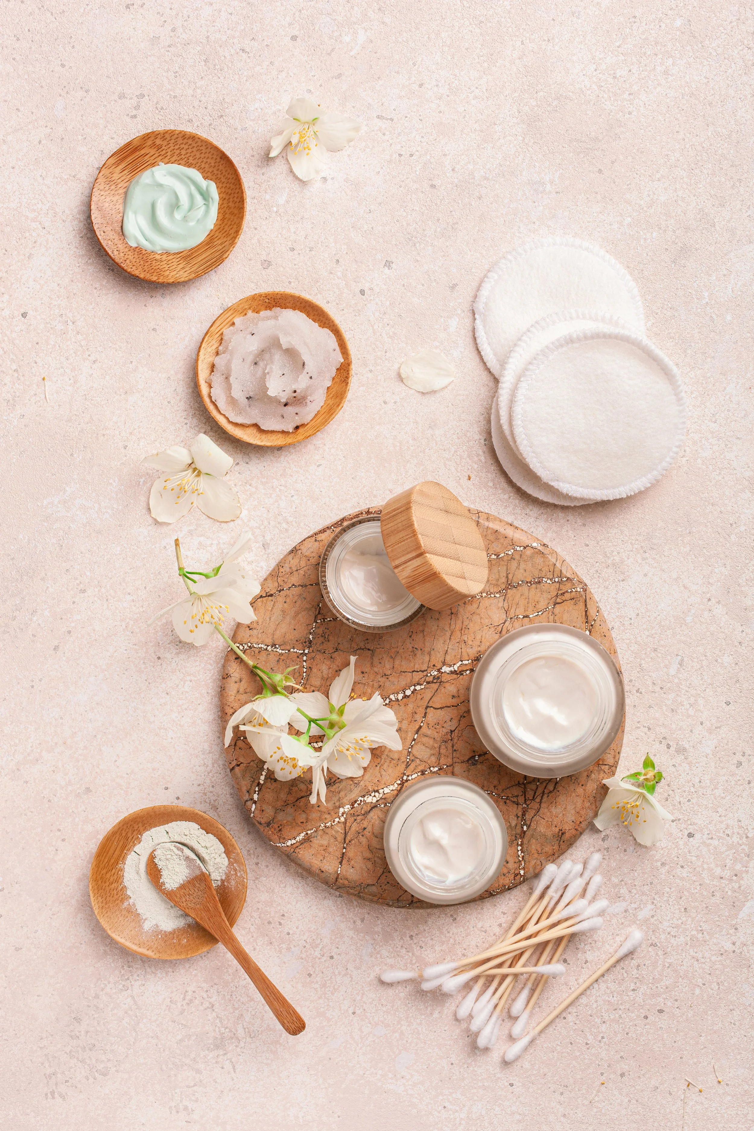 Collection of skincare products including creams and lotions in glass jars, cotton swabs, and cotton pads arranged on a marble and beige textured surface with white flowers around.