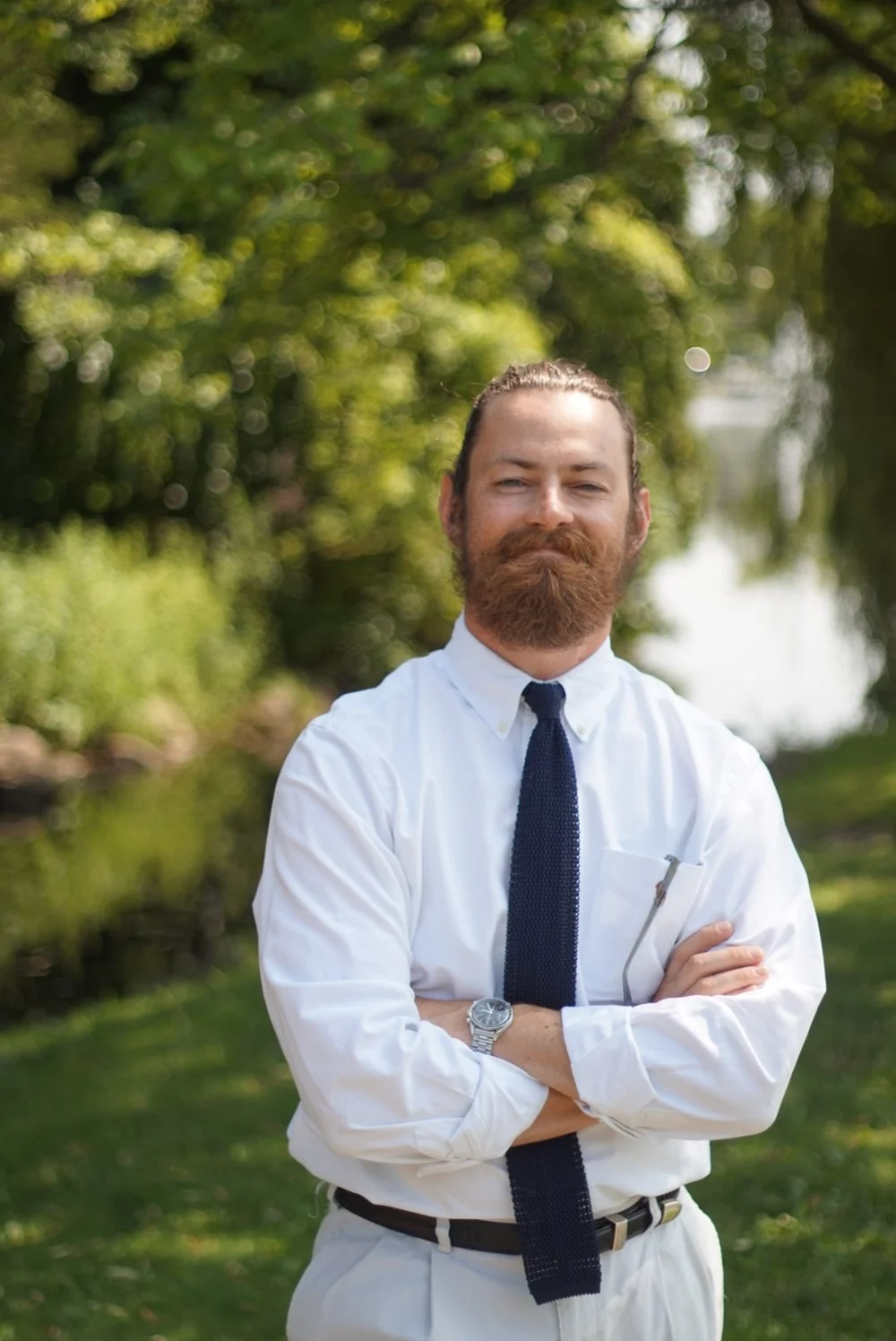 Kyle Porro in a white shirt and navy tie, arms crossed, in a park