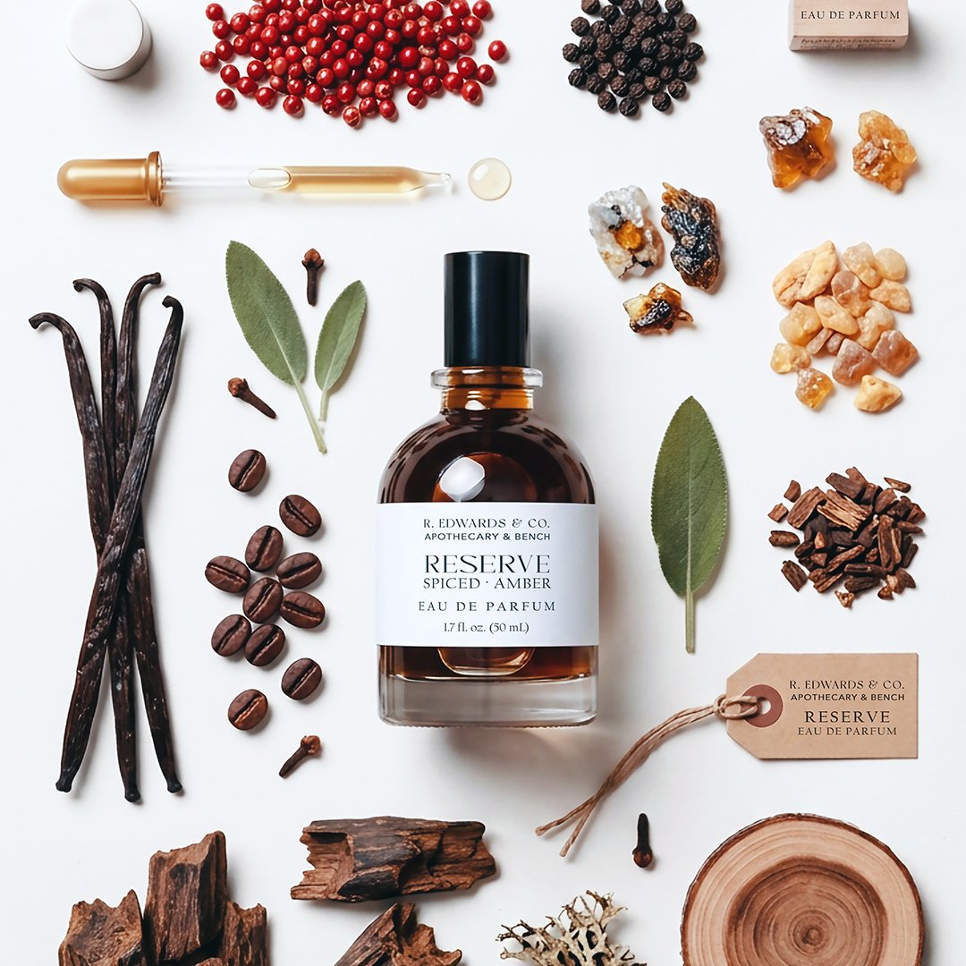 A collection of aromatic ingredients and a bottle of perfume labeled 'Reserve Spiced Amber Eau de Parfum' on a white background. Items include dried vanilla beans, coffee beans, various rocks and minerals, sage and bay leaves, pieces of wood, and a small perfume tag.