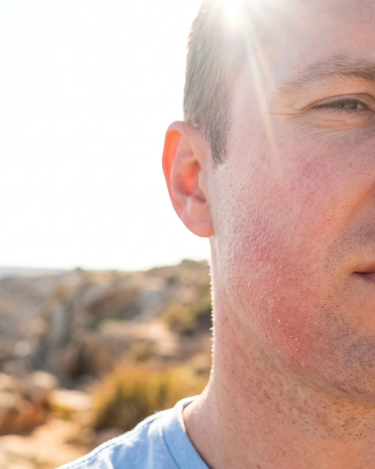 Close-up of a young man's face, showing his eye, ear, and part of his cheek, with sunlight in the background and an outdoor landscape.